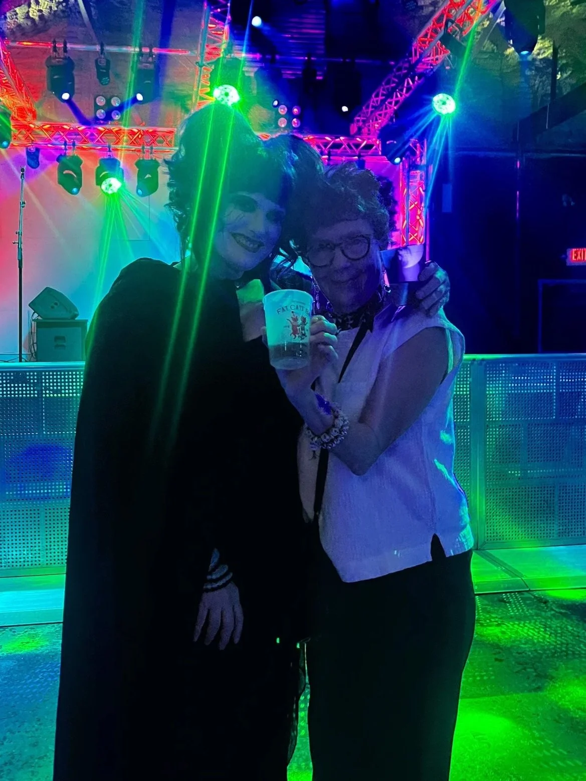 Two women smiling and holding a drink, standing in a colorful stage with bright lights, speakers, and a metal dance floor at a party or concert.