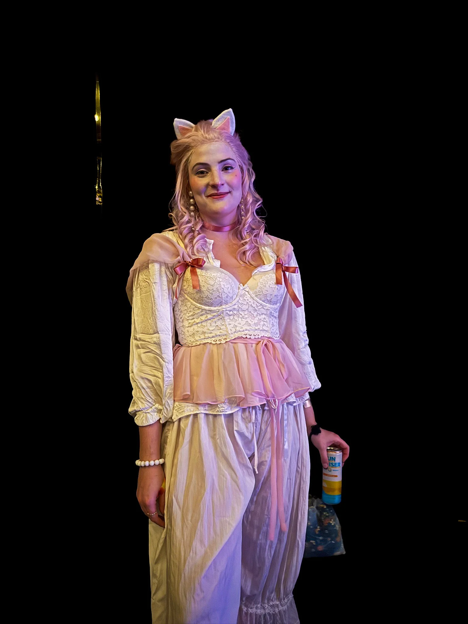 A woman dressed as a cat with pink bunny ears, wearing a vintage-style yellow dress with pink ribbons, standing against a black background. She has pink curly hair, pearl earrings, and is holding a can of disinfectant spray.