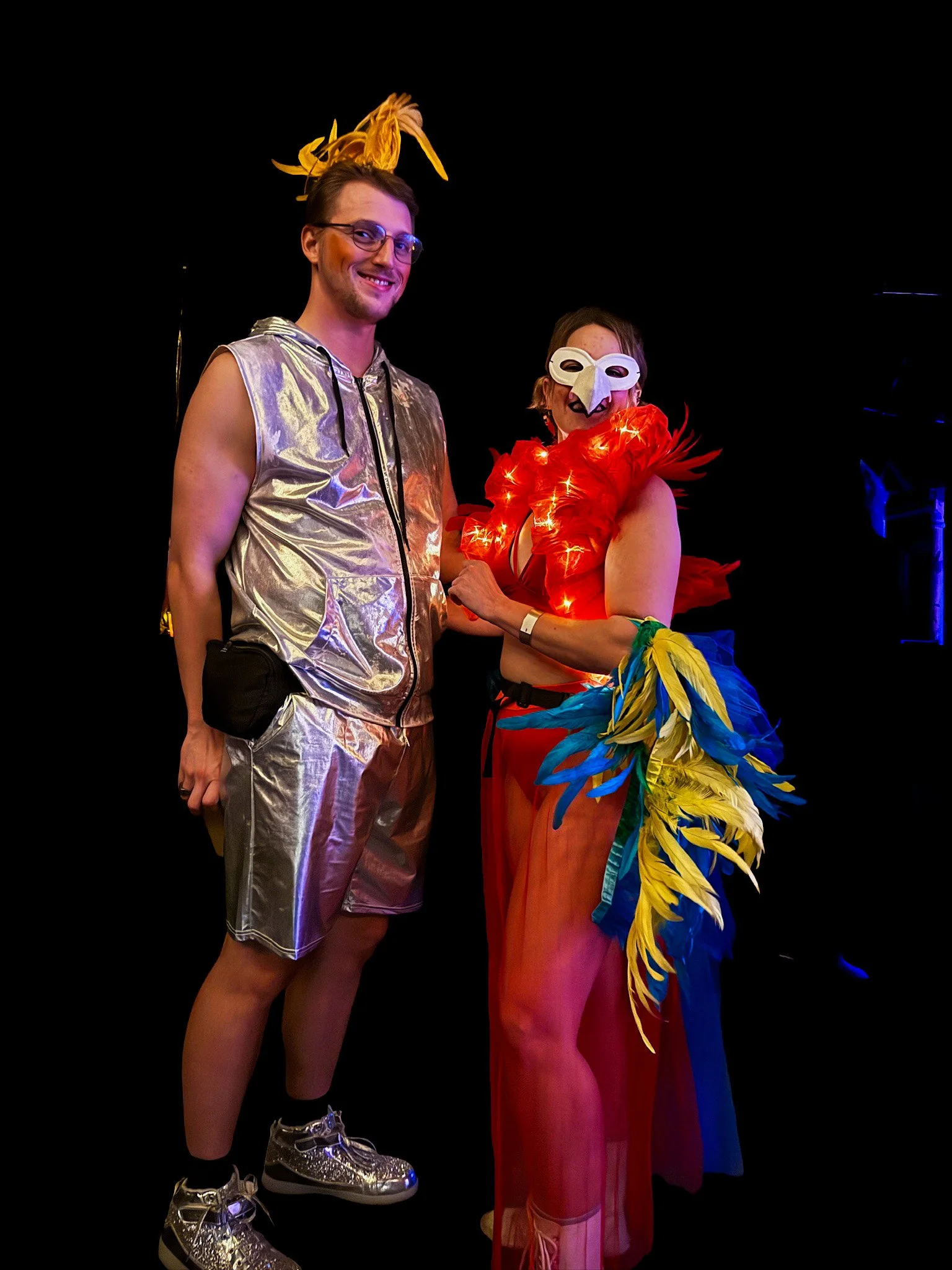 Two people dressed in colorful, theatrical costumes, standing against a black background. The person on the left wears a shiny silver sleeveless hooded top and matching shorts with metallic sneakers and glasses, smiling at the camera. The person on t