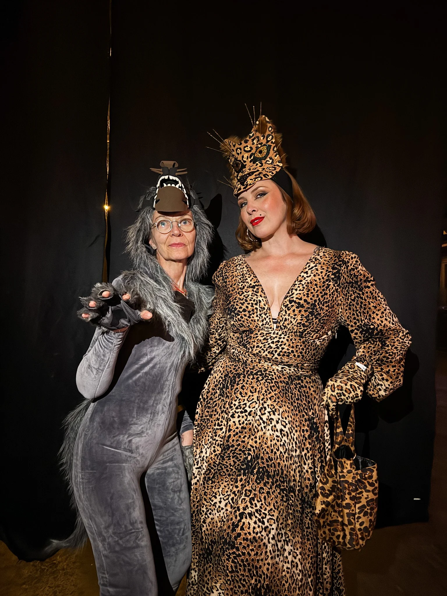 Two women dressed in animal costume outfits, one as a wolf and the other as a leopard, standing against a dark background.