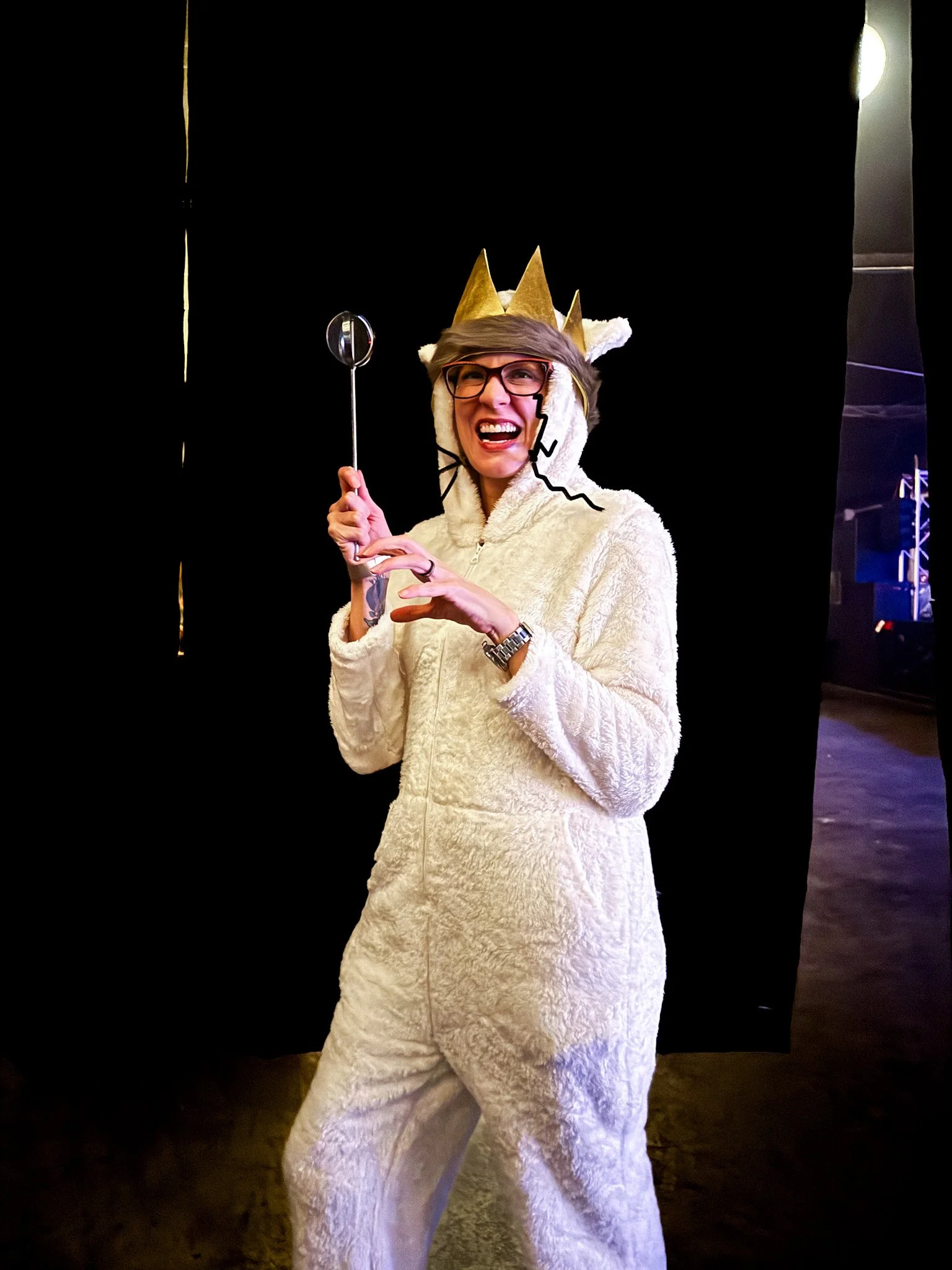 Person wearing a cream-colored fuzzy onesie, glasses, and a paper crown, holding a ladle and smiling, with a black backdrop and stage lighting in the background.