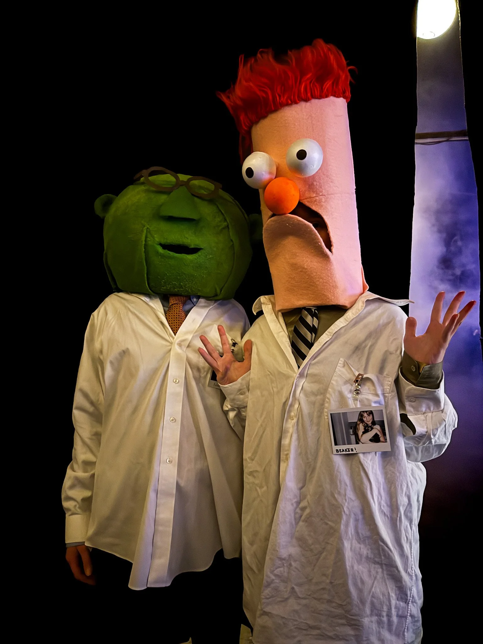 Two people wearing costumes with oversized head masks, one resembling a green head with glasses, and the other resembling a pink head with red hair, large eyes, and a clown nose, dressed in white shirts and ties, standing against a dark background.