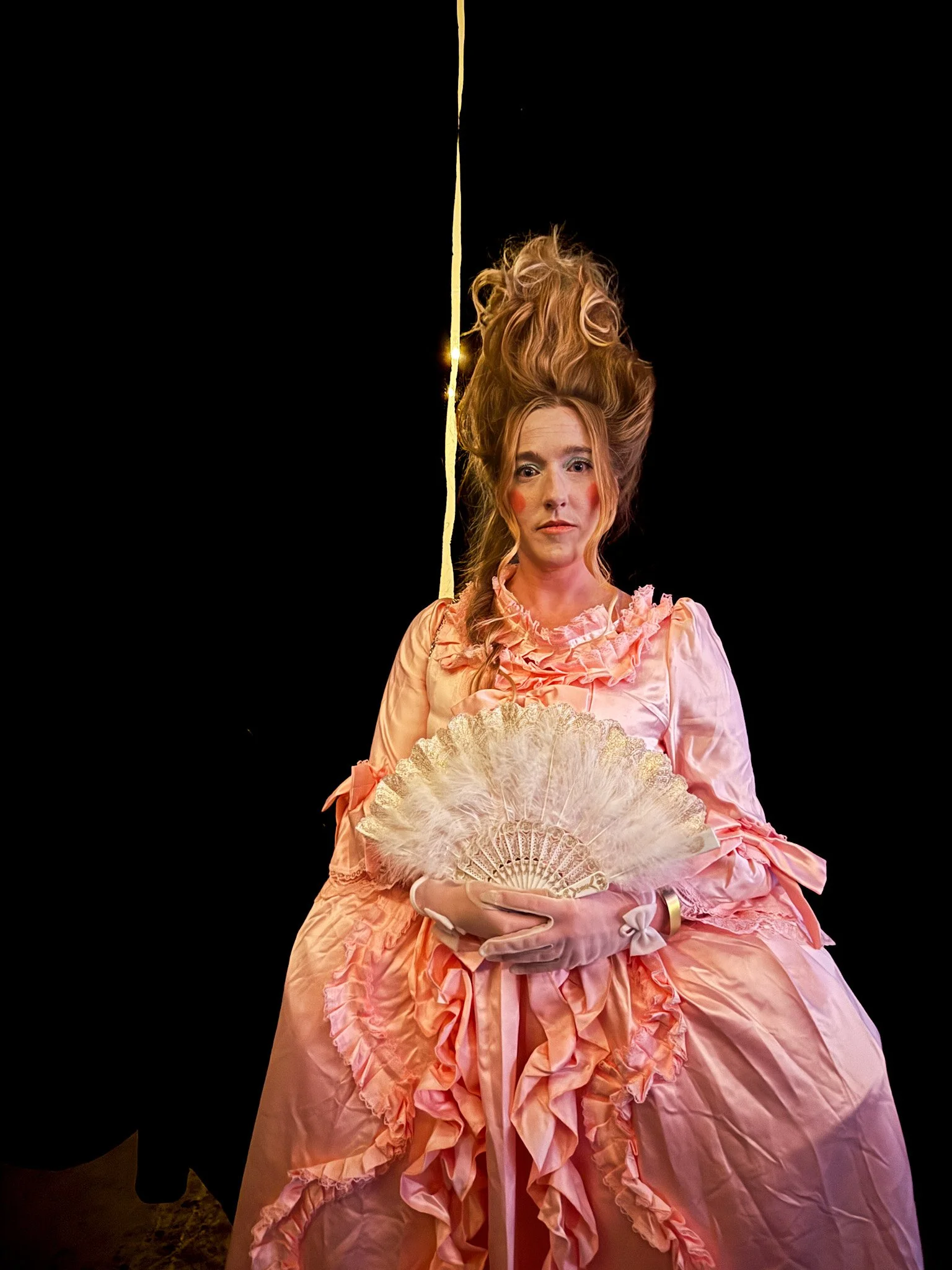 Woman dressed in pink vintage-style dress with ruffles, holding a lace fan, with elaborate updo hairstyle, standing against a black background.