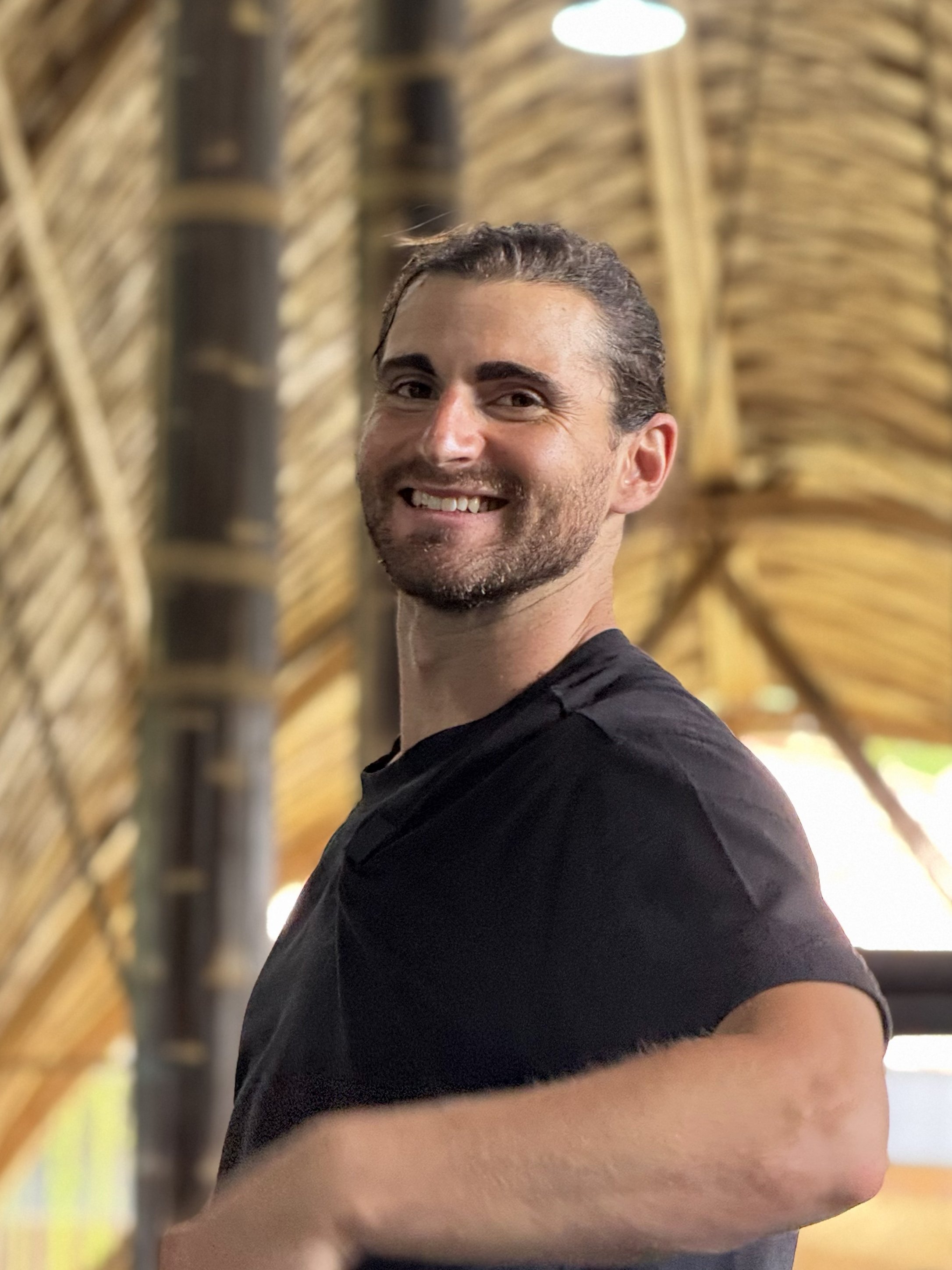 A smiling man with short dark hair and a beard stands in front of a wall made of woven wood with warm lighting.