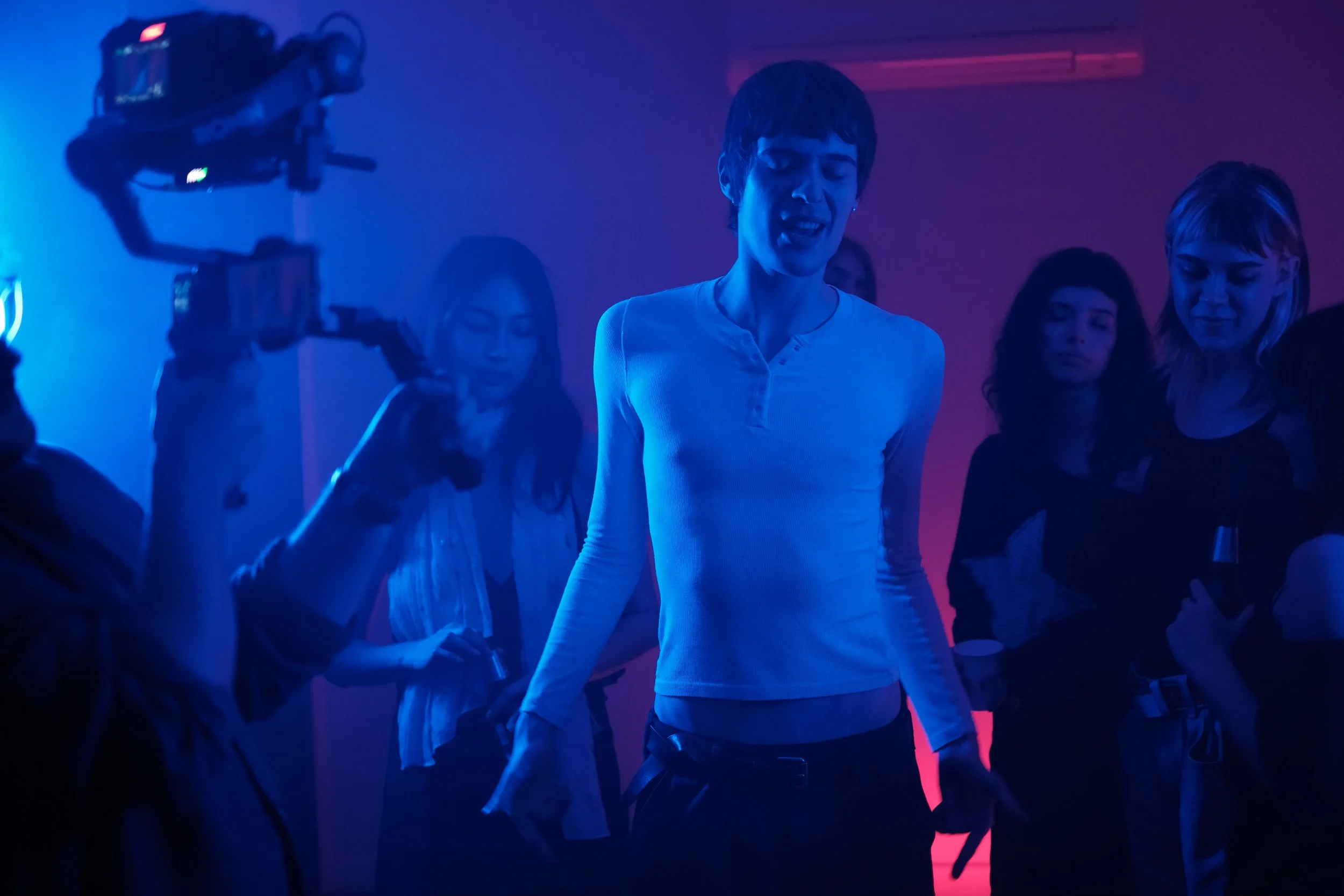 Group of women socializing at a party with pink and blue lighting, one woman in the center appears to be singing or speaking passionately.