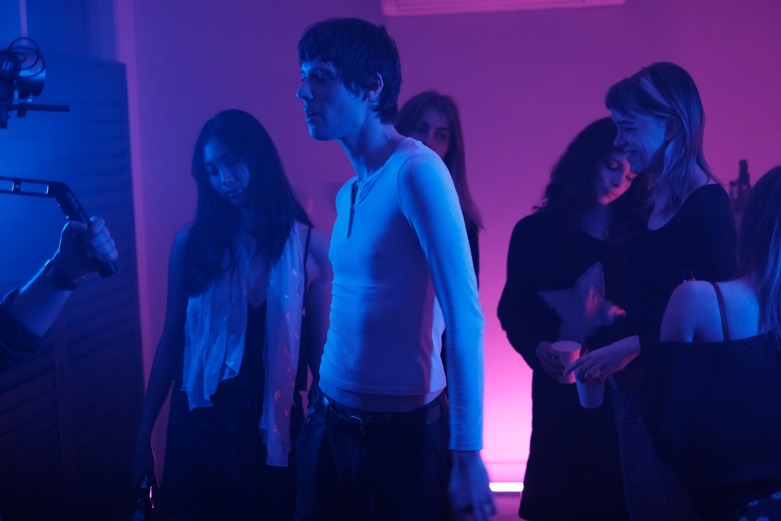 Group of young people at a party under purple and pink lighting, some holding cups, with a person on the left filming them with a handheld device.