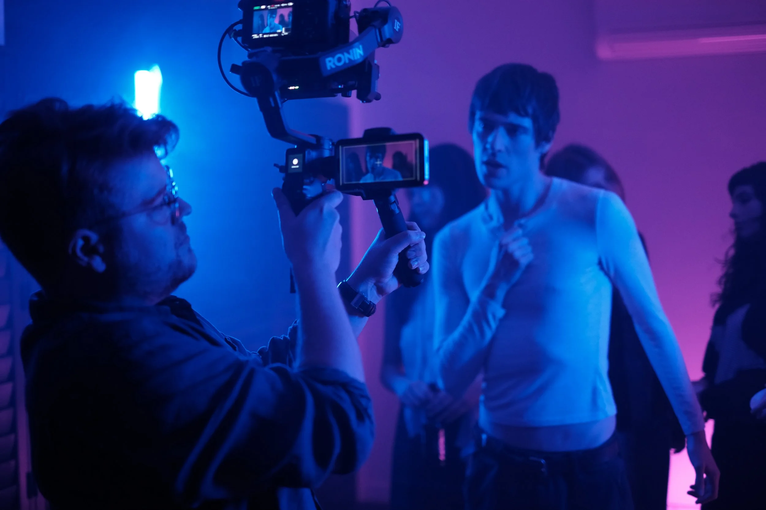 A person filming with a camera on a gimbal captures a woman in a white top at a social event with purple and blue lighting.
