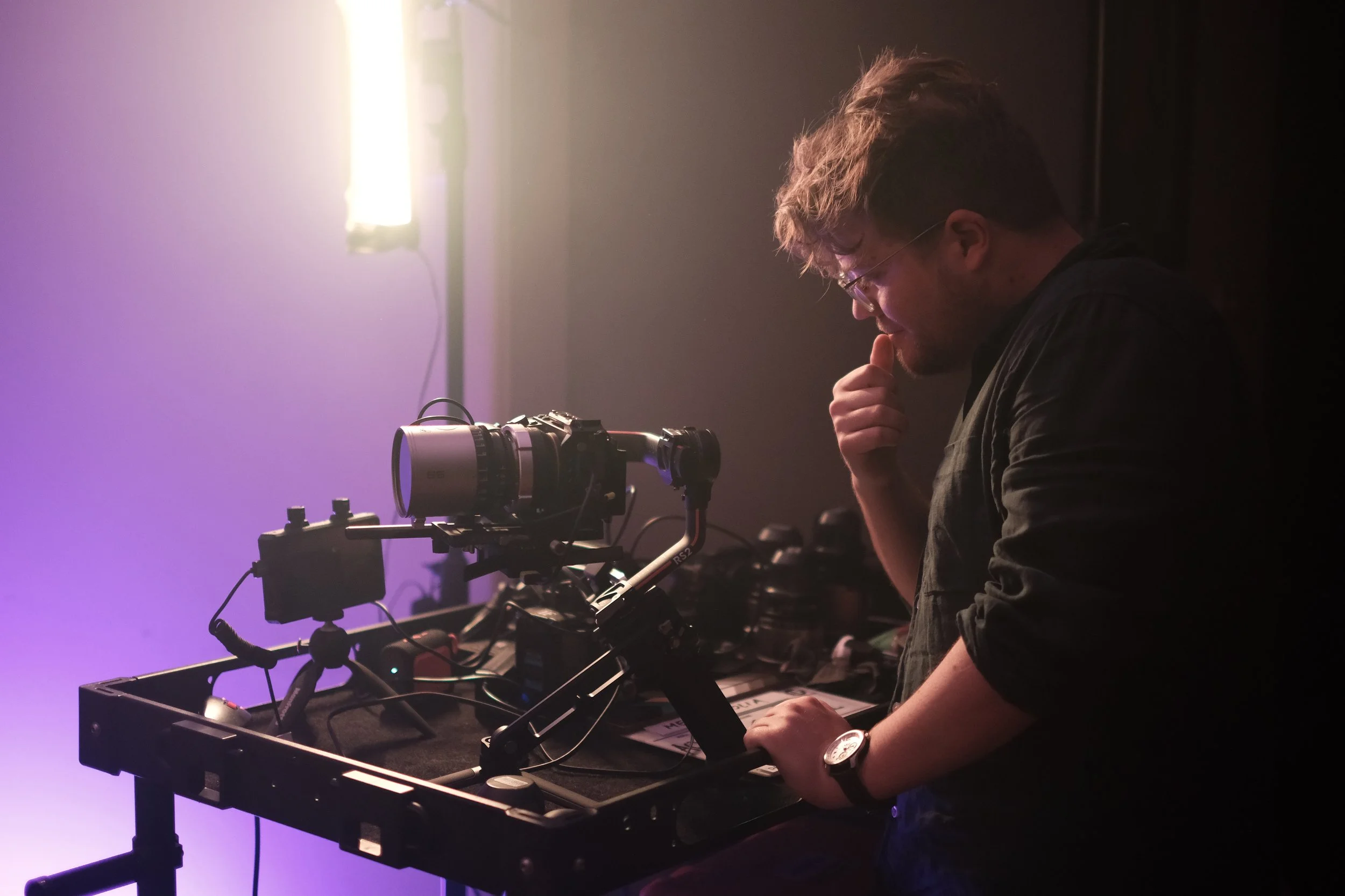 A person with glasses and a watch looking thoughtfully at a camera setup on a table with various equipment, illuminated by bright lighting in a dark room.