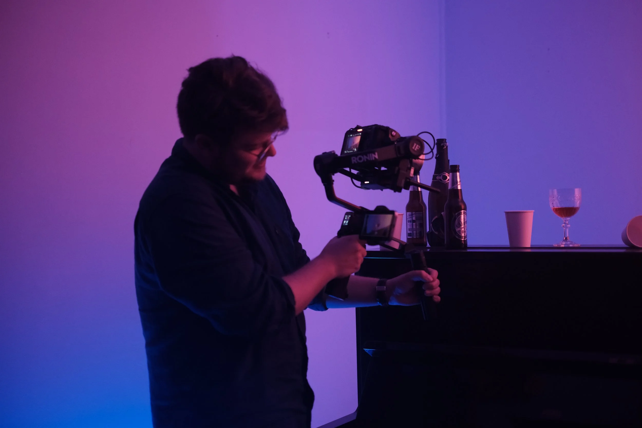 Person filming with a camera in a dimly lit room with bottles and drinks on a table.