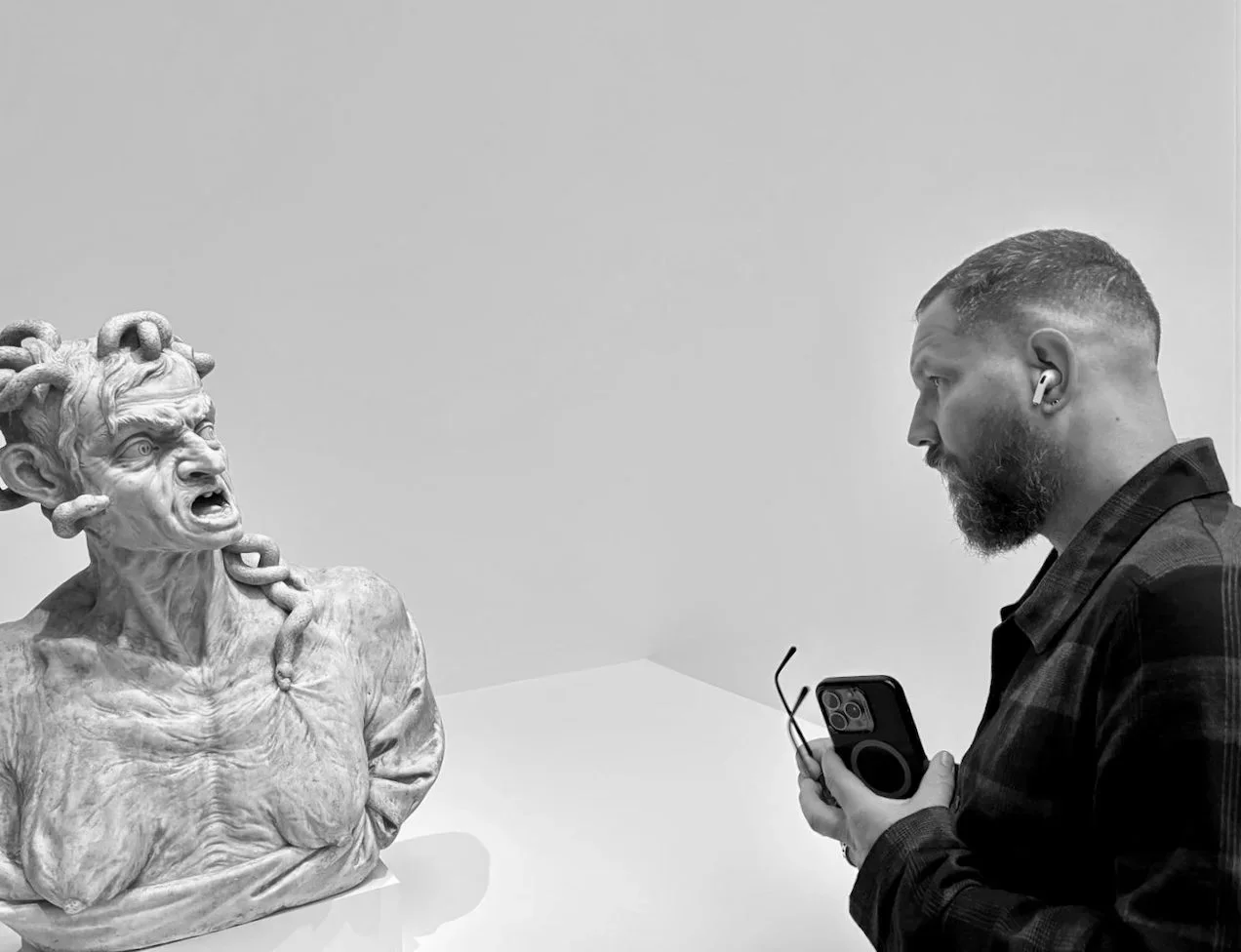 Artist Renzo Brandsma, bearded and wearing an earbud, holds his phone while looking at a sculpture of Medusa with snake-covered hair in a museum.