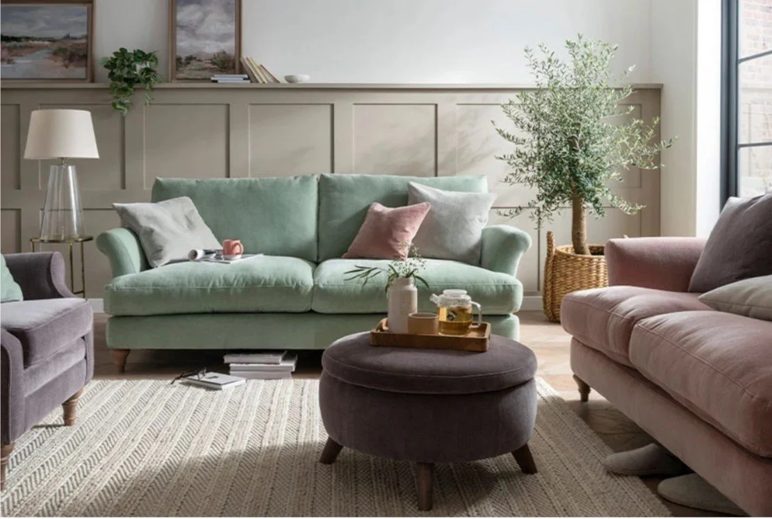 Living room with pastel green sofa, pink and white cushions, a purple armchair, a pink sofa, a round ottoman with a tray, and a tall potted plant near a window with natural light.