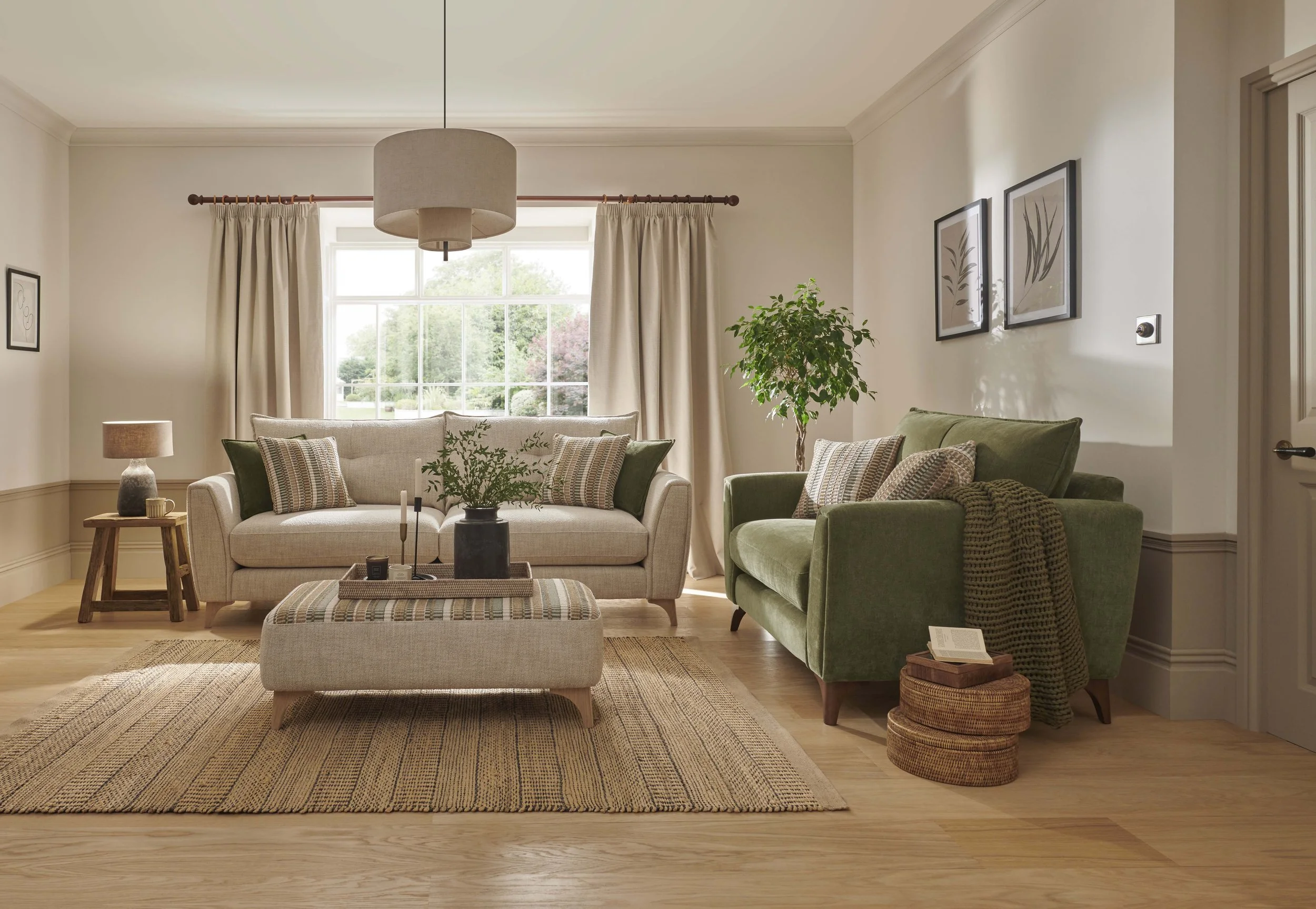 Living room with beige and green furniture, a large window with curtains, indoor plants, framed wall art, and a hanging ceiling light.