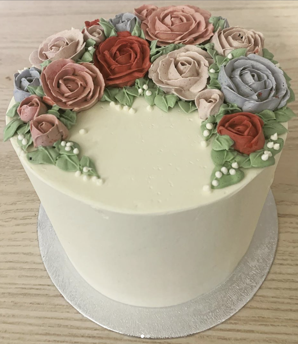 A round cake decorated with pastel-colored buttercream roses and green leaves on top, featuring small white decorative beads.