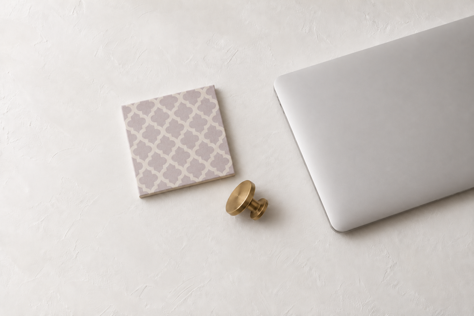 Corner of a silver laptop, a brass drawer pull, and a lavender Moroccan-pattern tile on a white plaster surface.