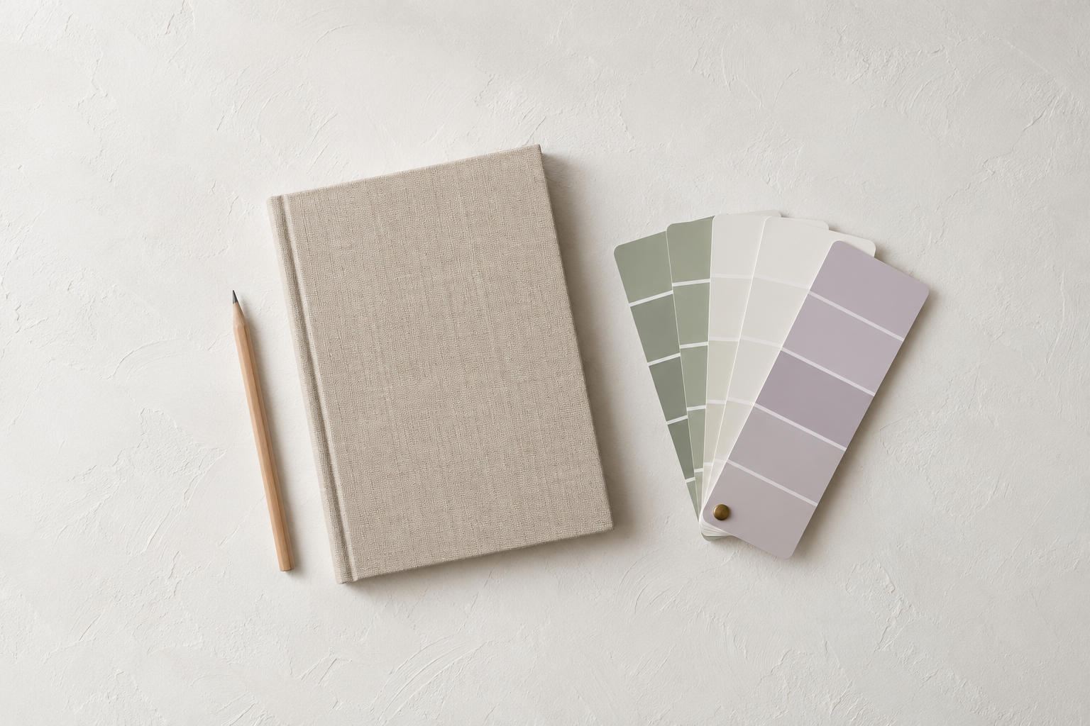 Linen notebook, pencil, and a fanned paint swatch deck in sage, white, and lavender on a white plaster surface.