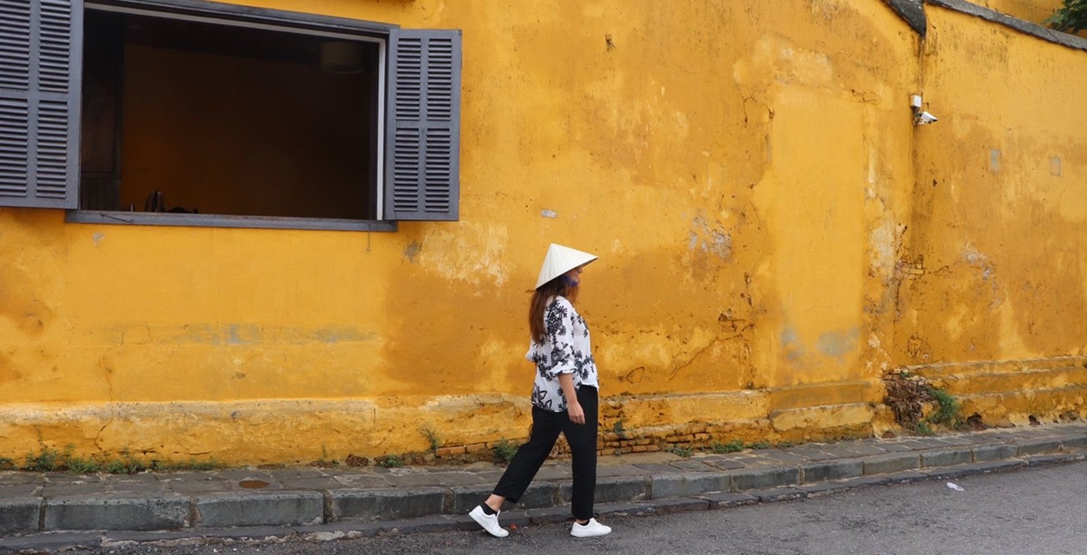 Woman wearing a traditional Vietnamese conical hat walking past a yellow colonial wall in Hoi An