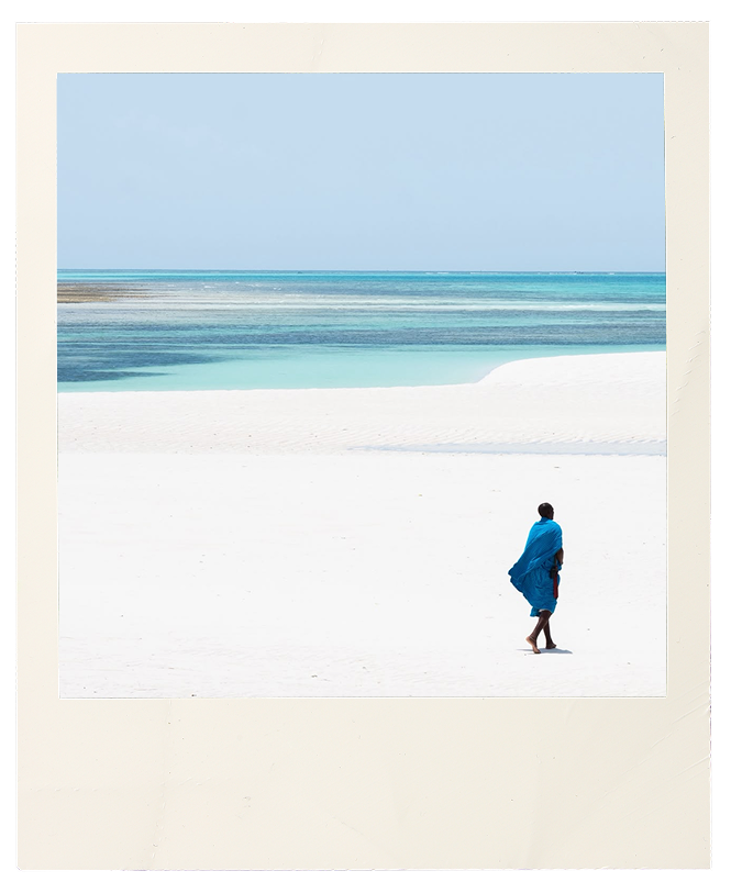 Person wrapped in a blue cloth walking along a vast white sand beach with turquoise water