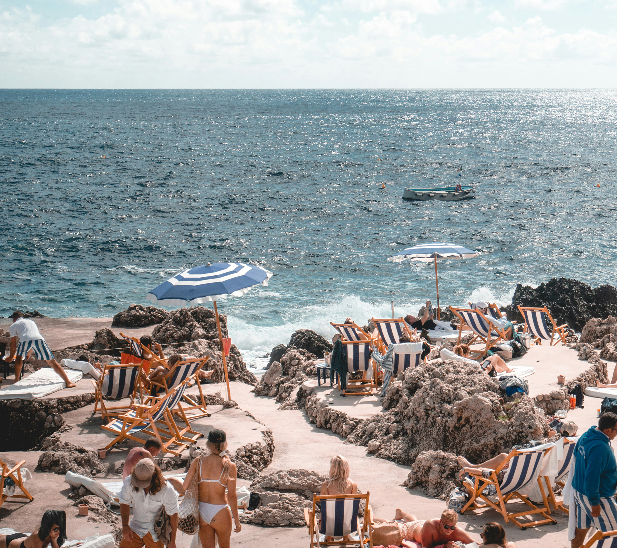 Guests relaxing on striped deck chairs at a rocky Mediterranean beach club terrace overlooking the ocean