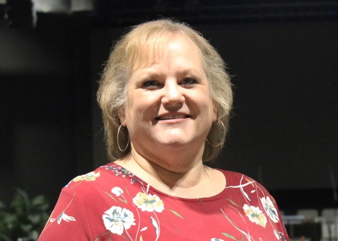 Smiling woman wearing a floral dress and hoop earrings, standing indoors against a dark background.