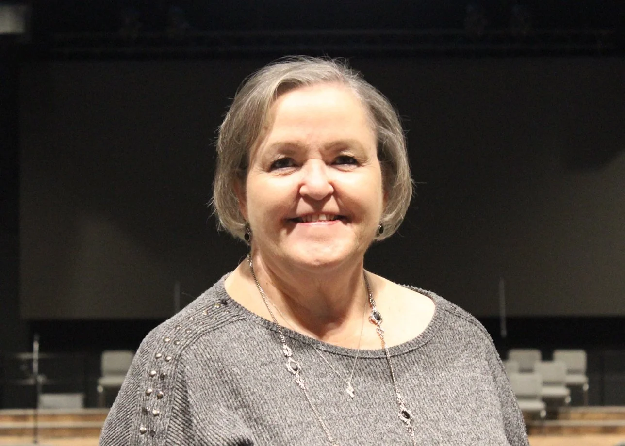 A person with short gray hair wearing a gray sweater standing indoors in front of a blurred background.