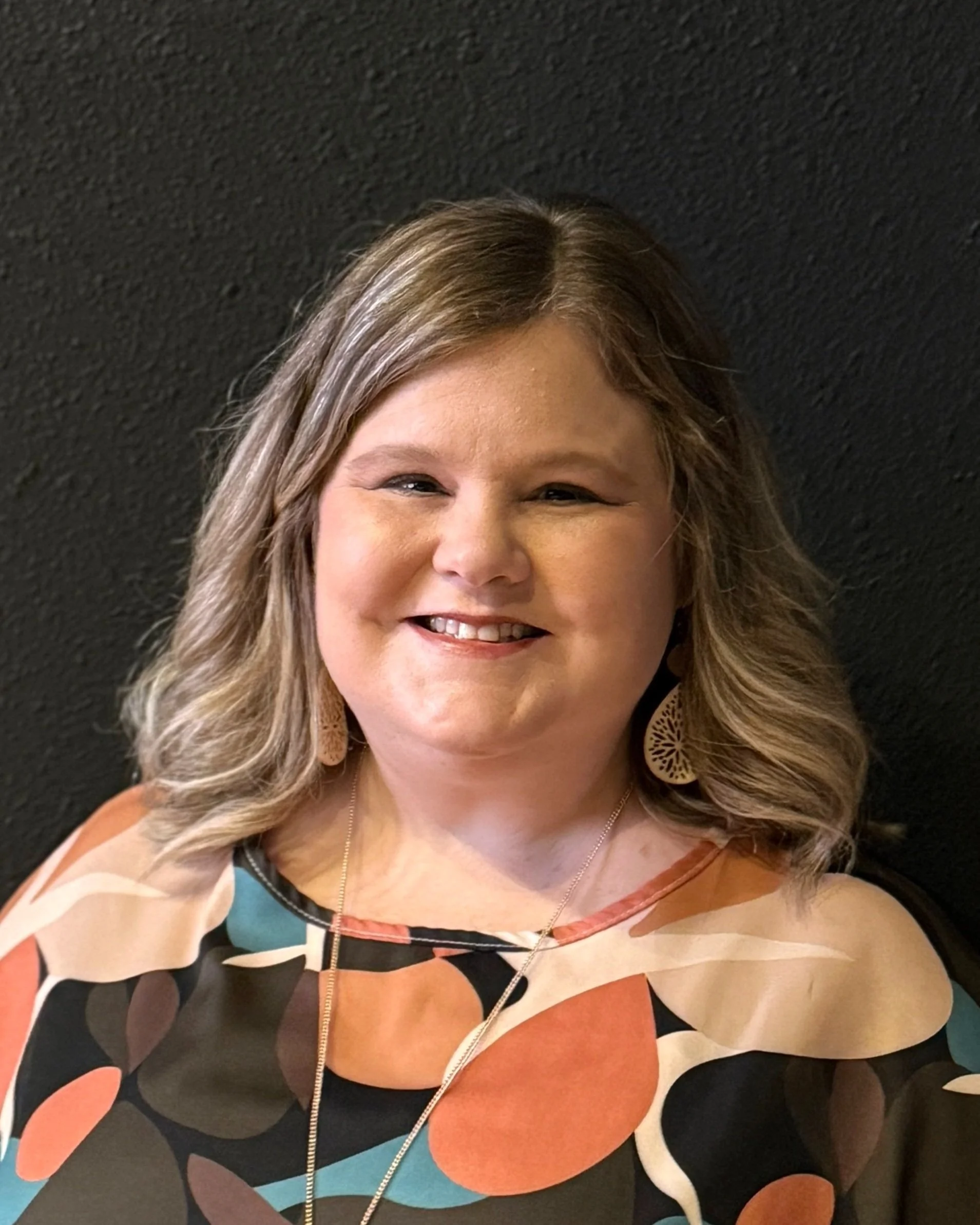 Smiling woman with shoulder-length blonde hair, wearing patterned top and large earrings, against a black background.