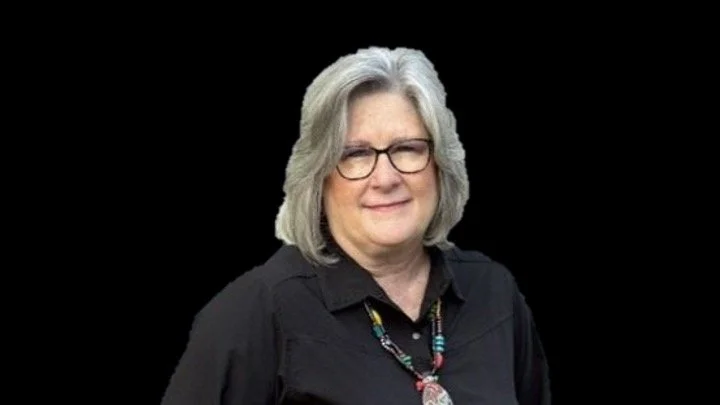 Woman with gray hair, wearing glasses and a black shirt with a colorful necklace, on a black background.