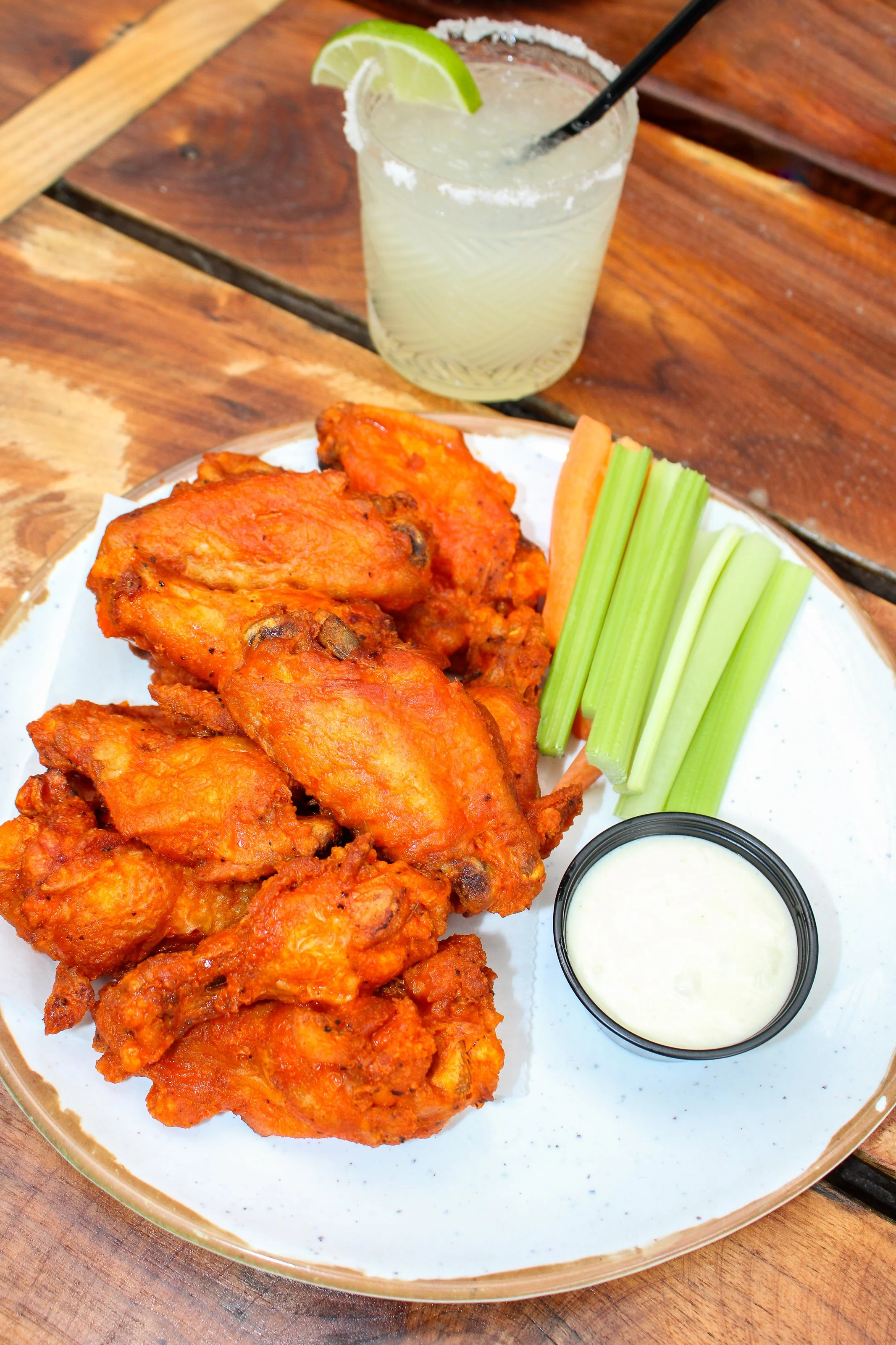 Plate of crispy buffalo-style chicken wings served with celery, carrots, and a side of blue cheese dressing on a wooden patio table, with a salted margarita garnished with lime in the background.