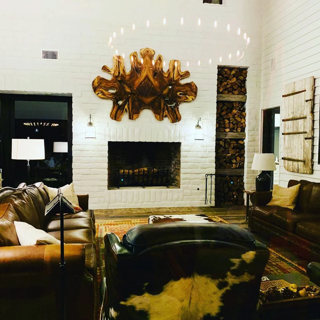 Cozy living room with brick fireplace, leather sofas, cowhide chair, and wood wall art. Stacked firewood decorates one wall, with candles creating ambient lighting.
