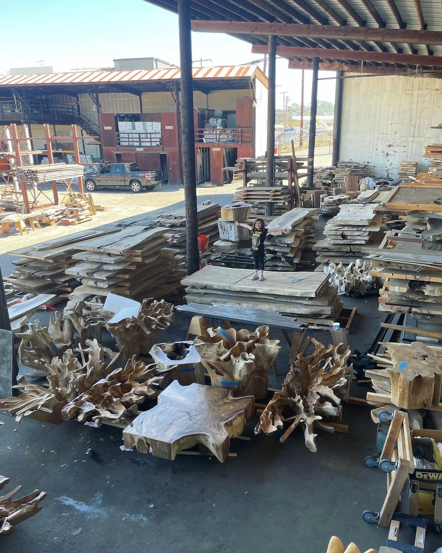 Outdoor storage area with stacks of wooden planks and raw wood pieces, including various thick slabs and unique, natural wood shapes. A metal roof covers part of the area. A person is standing among the wood pieces near the center.