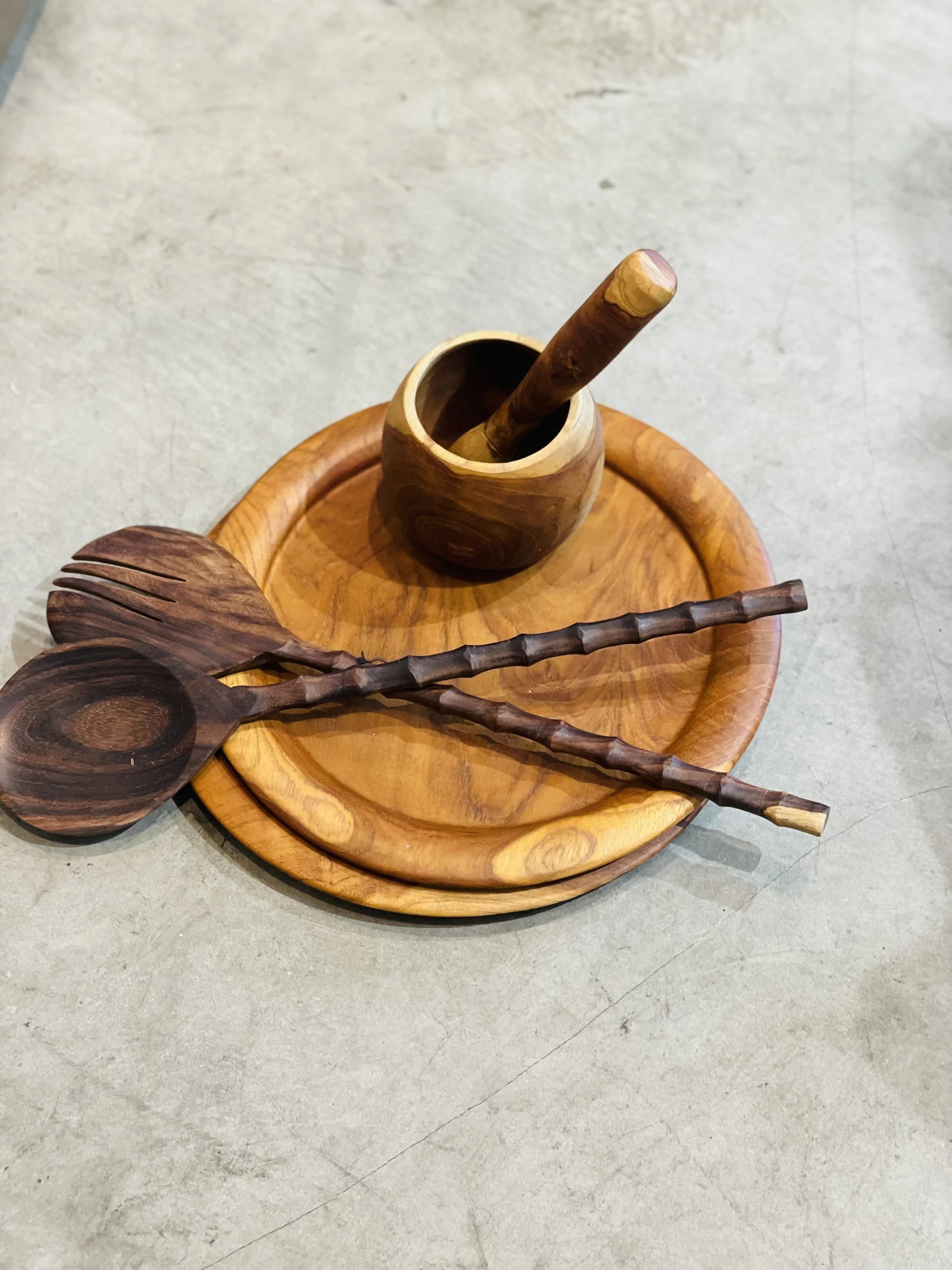 Wooden mortar and pestle with salad servers and plates on a stone surface.
