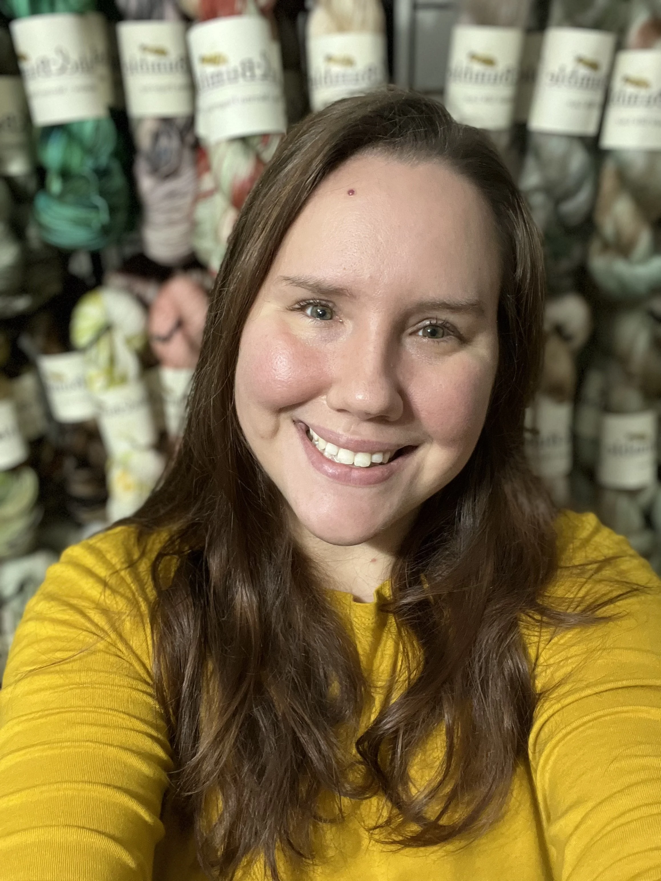A young woman with long brown hair and a yellow shirt smiling, standing in front of a wall of yarn skeins.