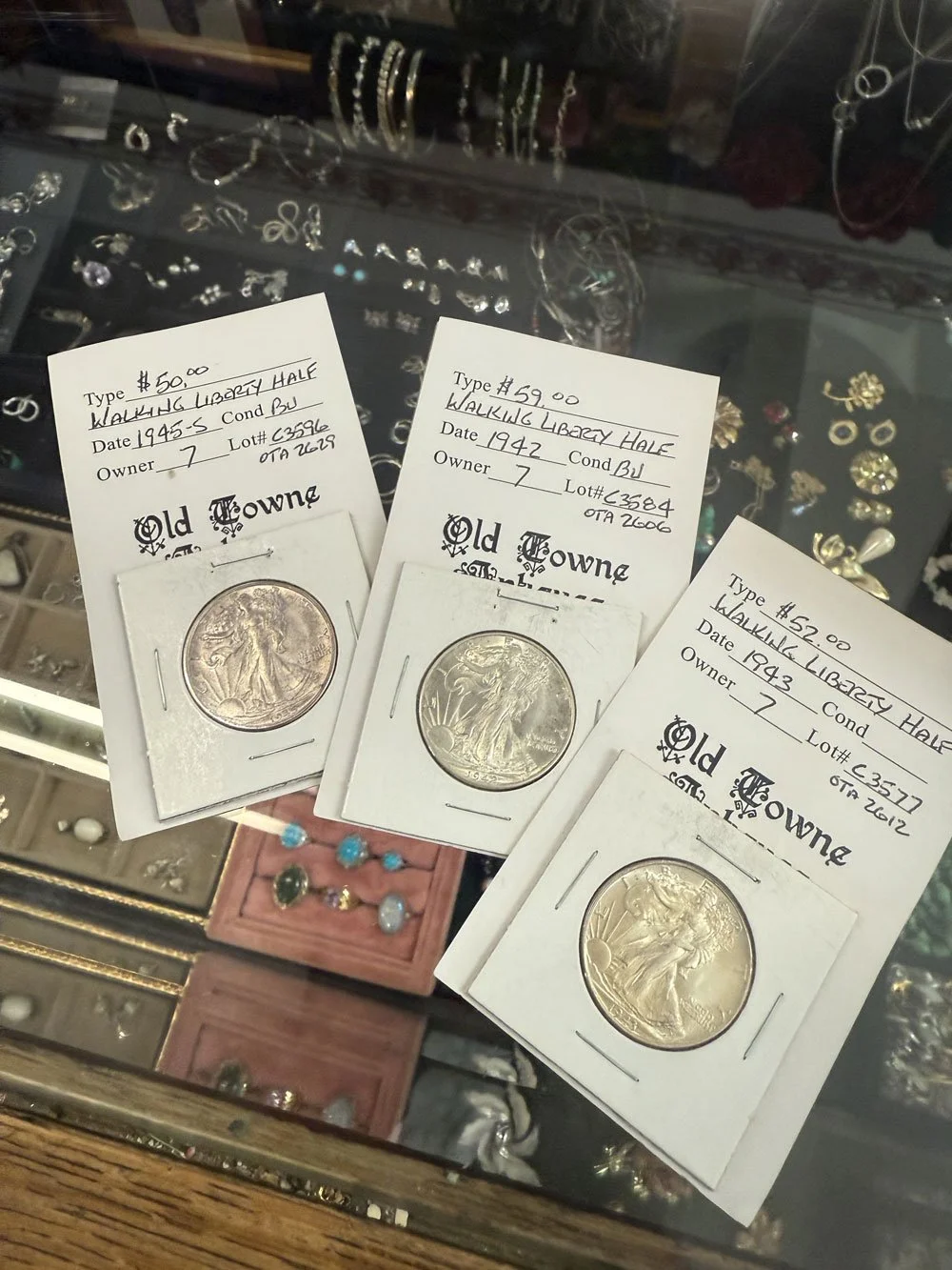 Three silver mercury dimes in protective holders with handwritten labels showing their prices, years, and lot numbers, displayed on a glass counter with jewelry in the background.