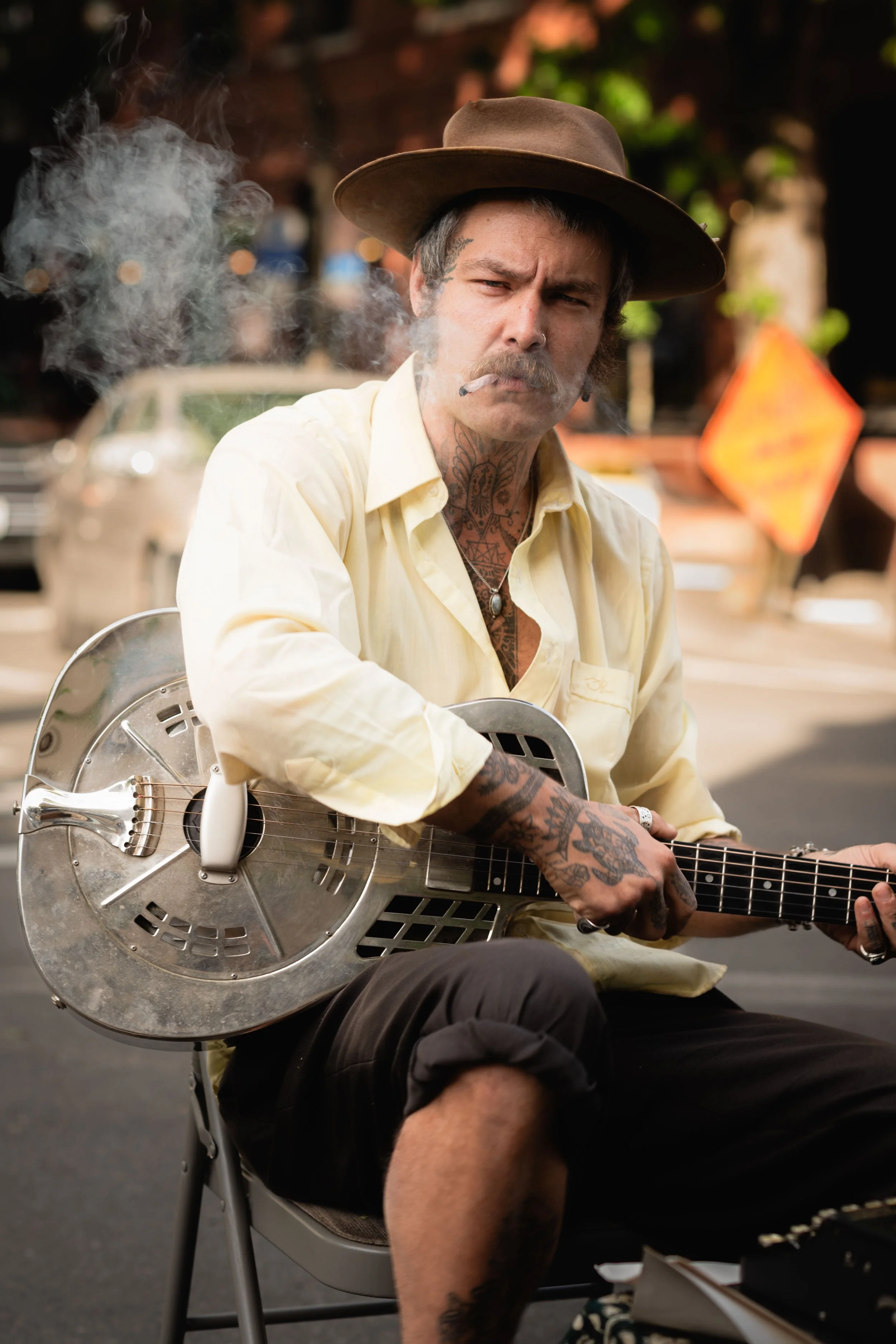 A man wearing a hat and a yellow shirt plays a resonator guitar while smoking in an outdoor urban setting.