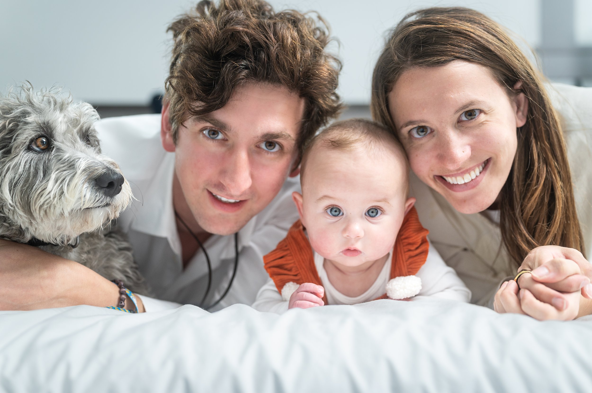 A family of three with a dog lying on bed, smiling and looking at the camera. The family includes a man, a woman, and a baby. The dog is furry and attentive.