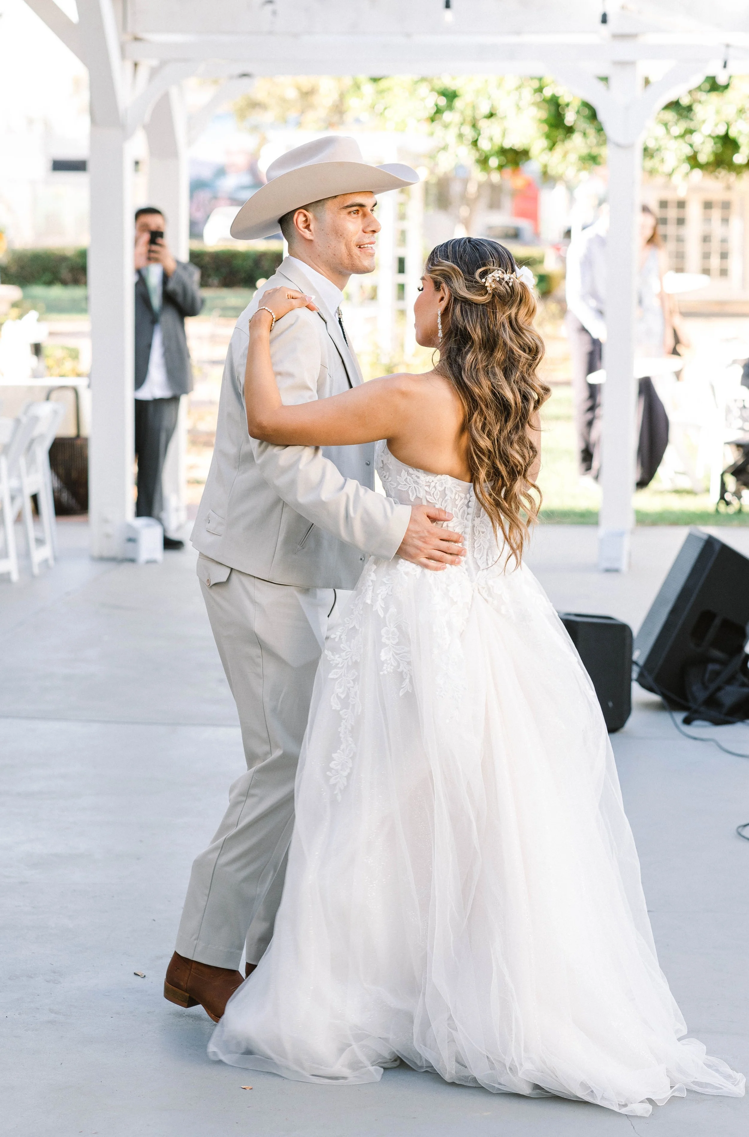 A bride and groom dance at their wedding reception outdoors with white wooden structures and guests in the background.