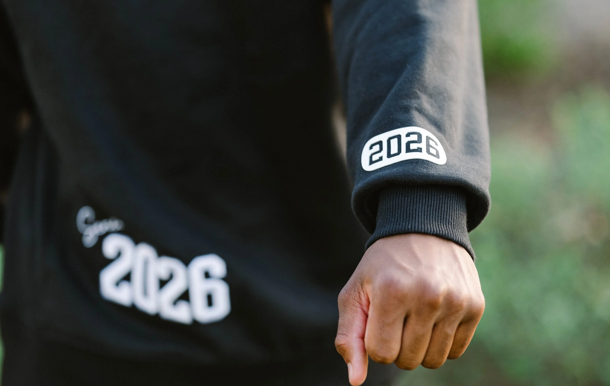 Close-up of a person's arm wearing a black hoodie with '2026' printed on the sleeve and a matching design on the pocket, with a clenched fist and visible veins, outdoors with a blurred natural background.