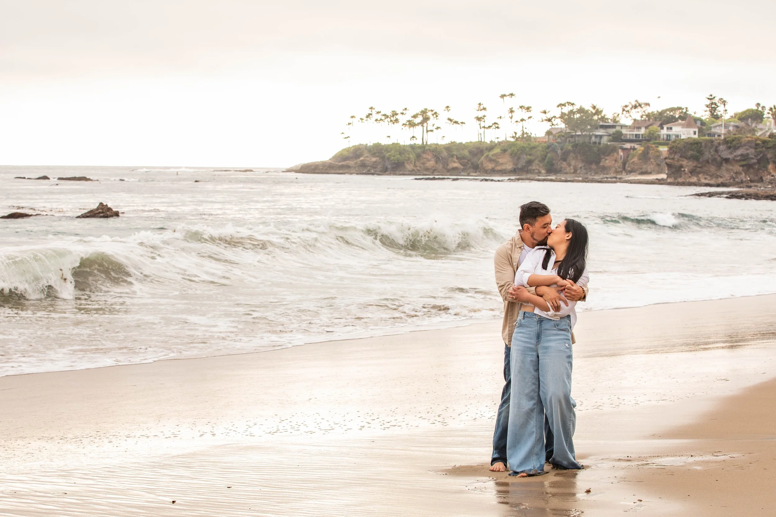 A couple kissing on a sandy beach with waves in the background and coastal cliffs lined with trees and houses.