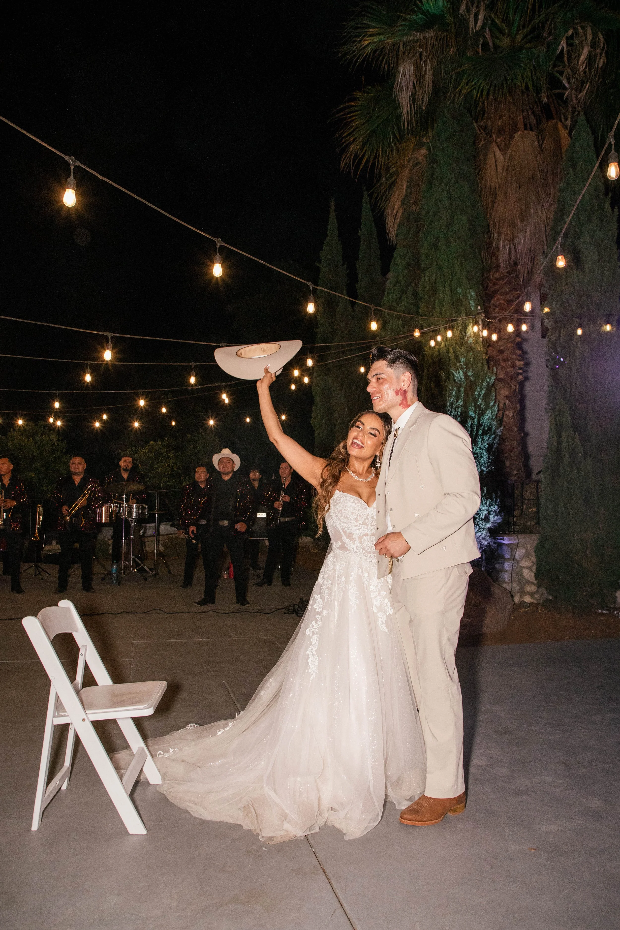 Couple, bride in white wedding dress and groom in beige suit, dancing at night outdoor wedding reception with string lights, band in background, palm trees, and chairs.
