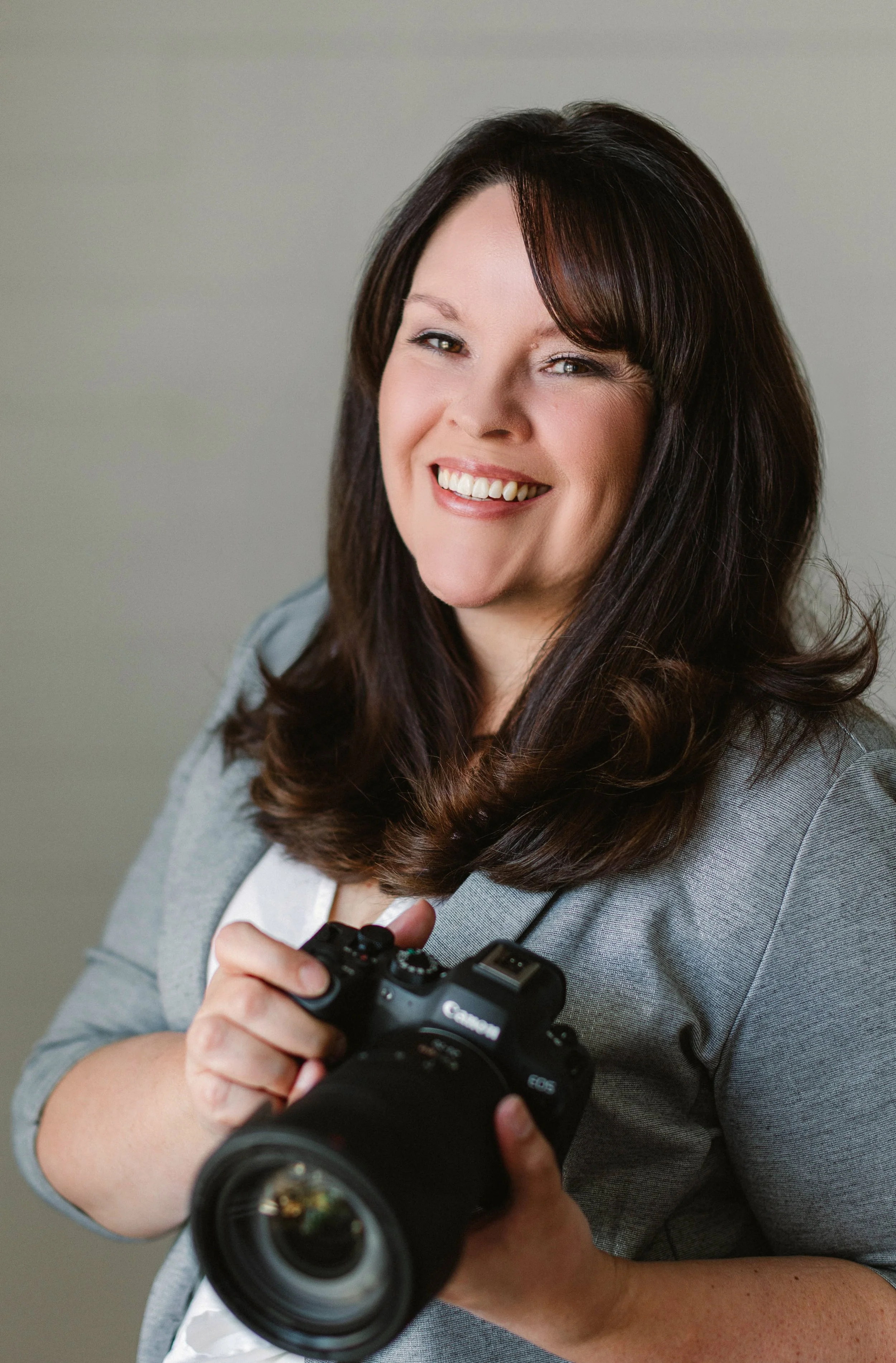 A woman with long dark hair smiling and holding a Canon DSLR camera.