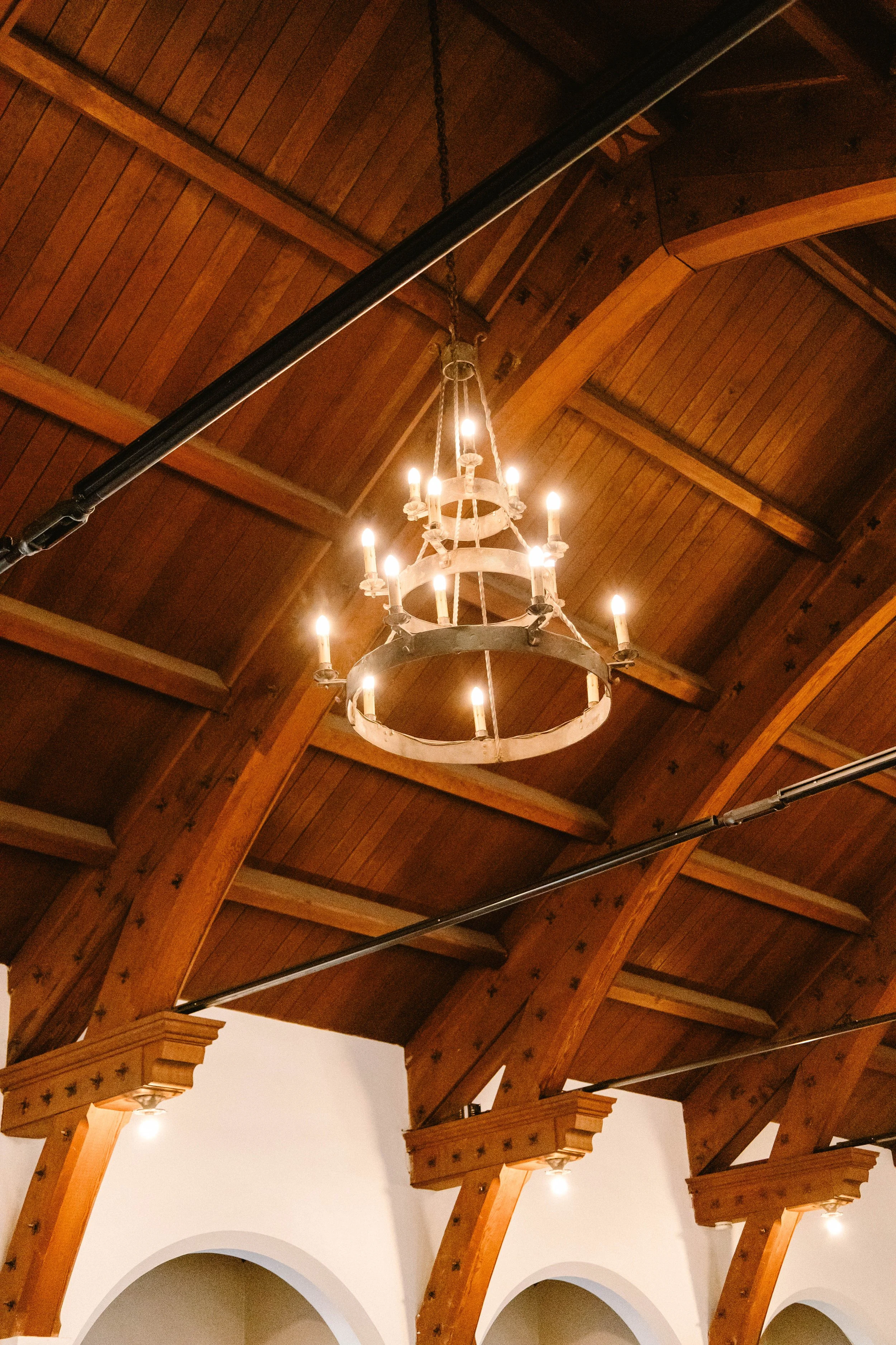 A chandelier with multiple lights hangs from a wooden ceiling, which has arched beams and decorative supports.