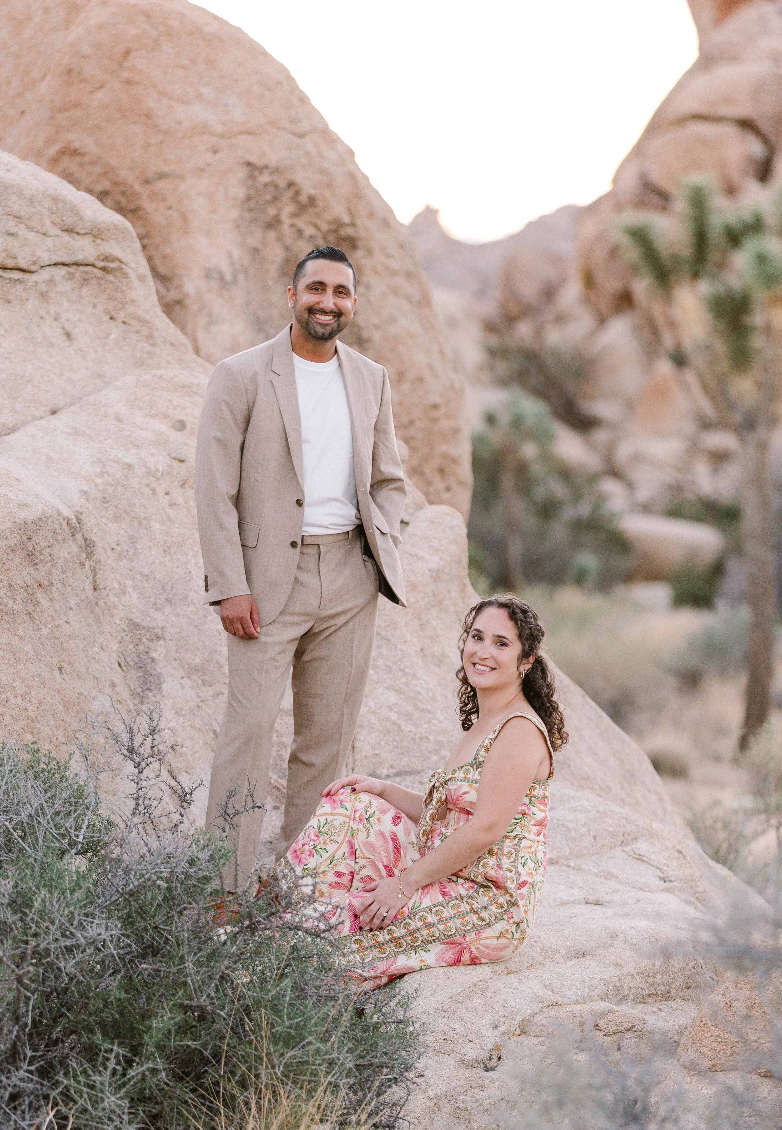 A man in a beige suit stands next to a woman in a floral dress sitting on a rock in a desert landscape with large boulders and sparse vegetation.