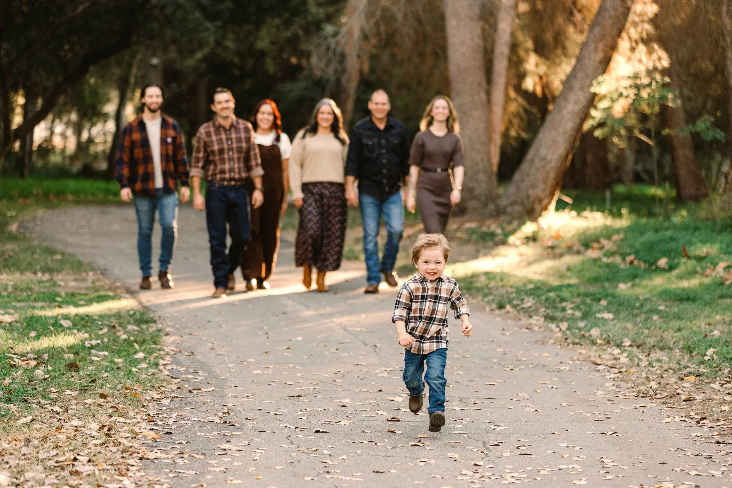 A young boy running happily in front of a group of adults walking along a park trail surrounded by trees and fallen leaves.