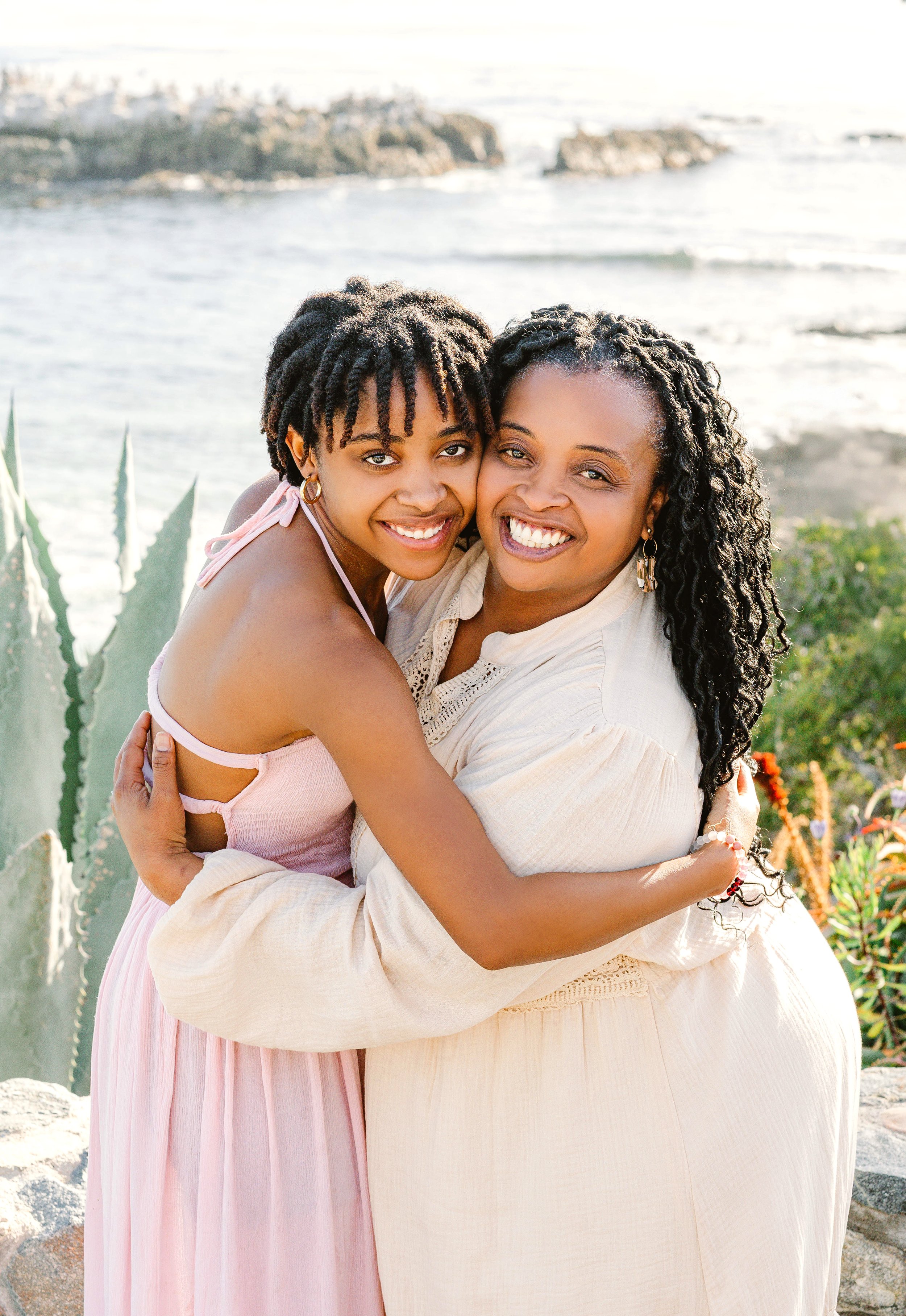 Two women hugging on a beach with waves and rocks in the background, smiling at the camera.