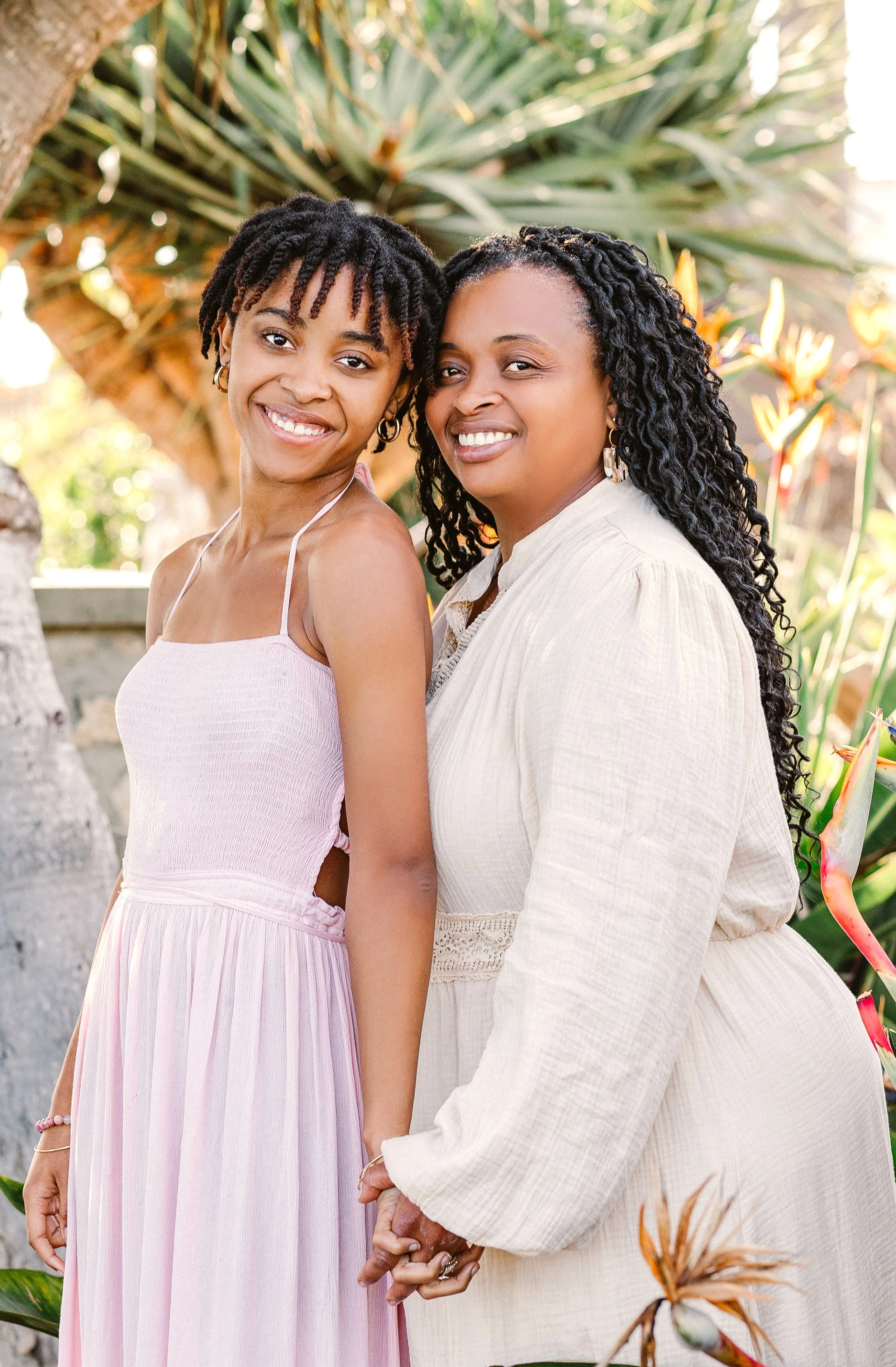 A young woman and an older woman standing outdoors, smiling and holding hands, surrounded by lush green plants.