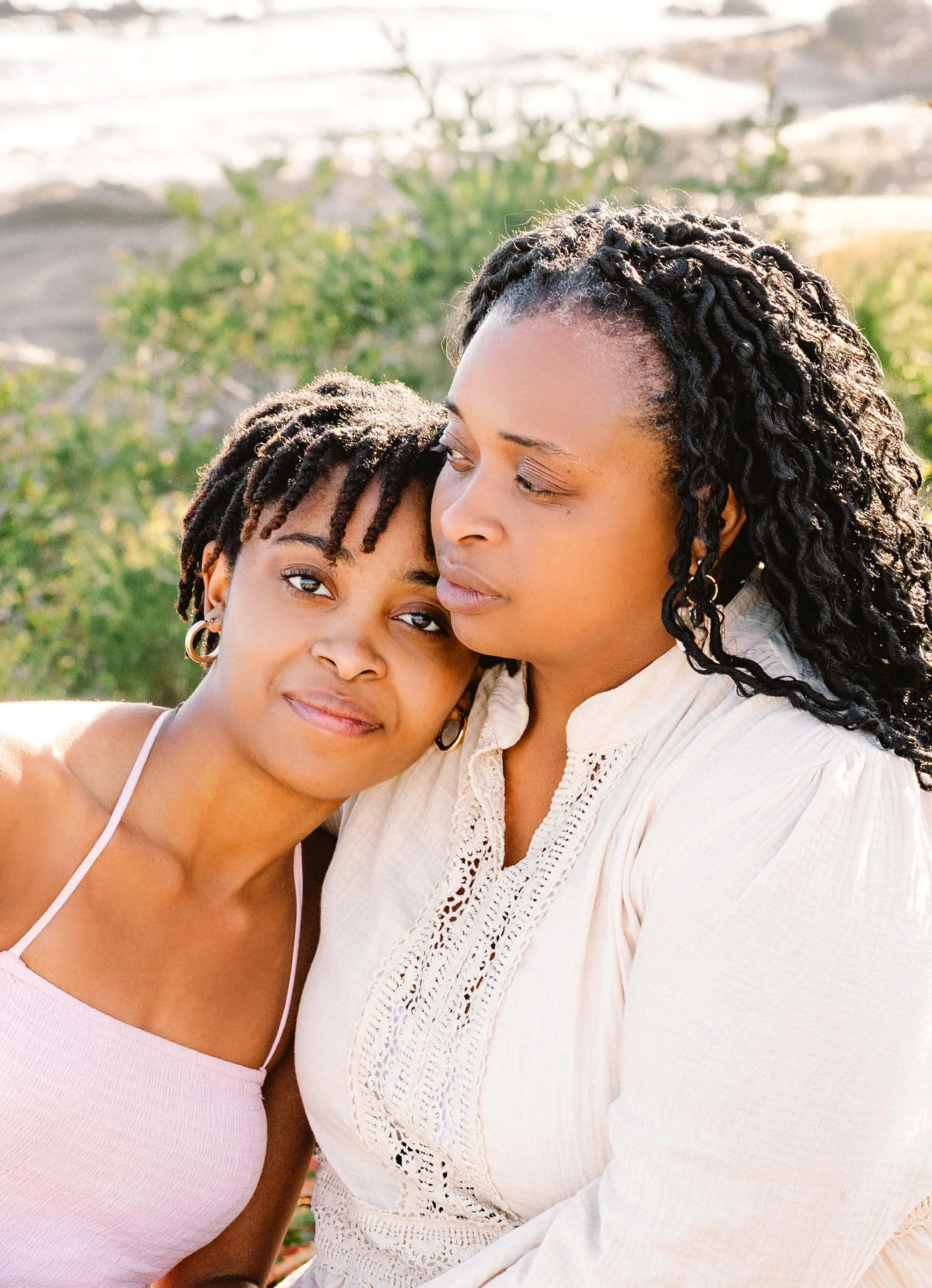 A woman and a girl with braided hair are close together outdoors, smiling softly at the camera with greenery in the background.