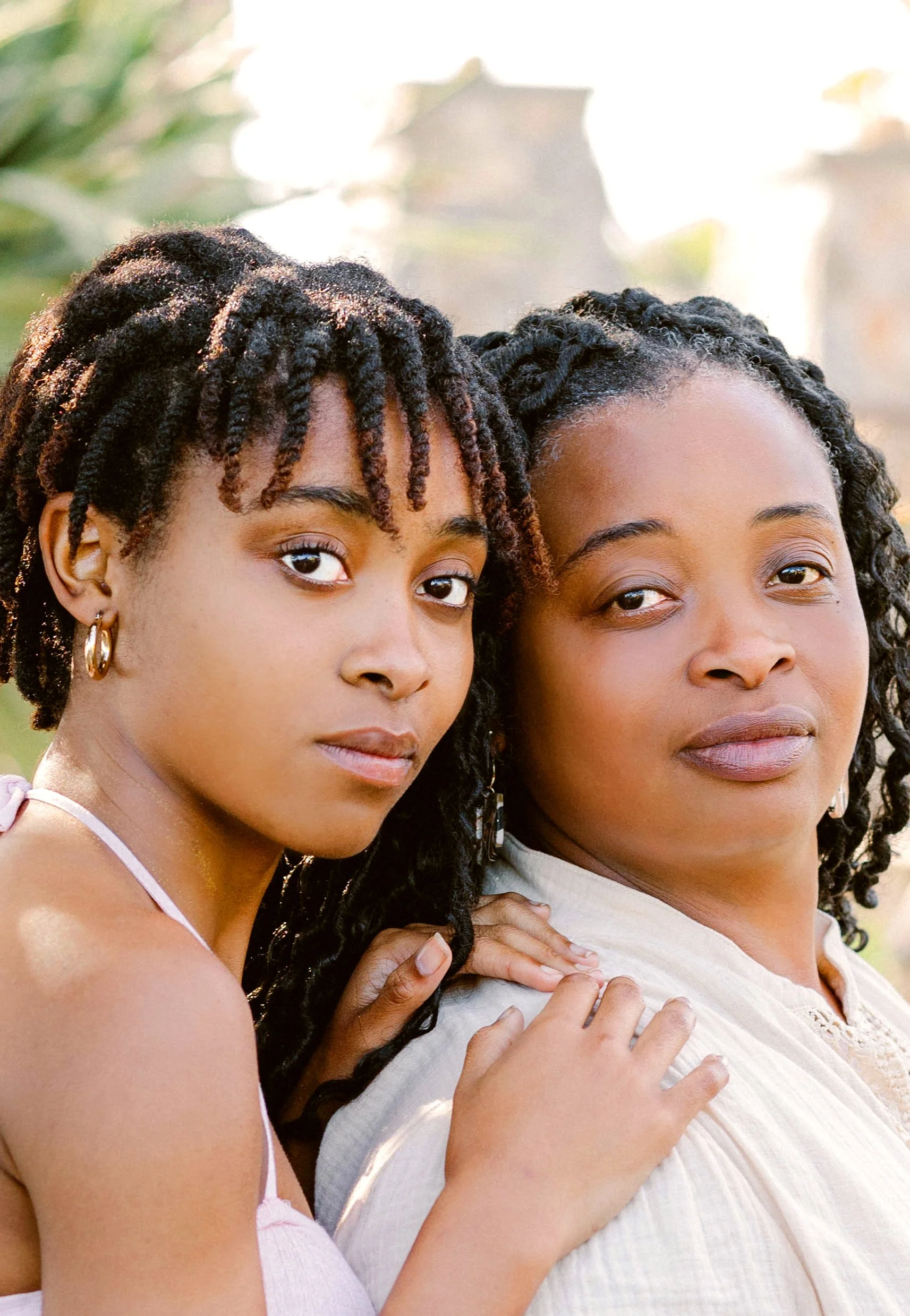 A young girl and woman with natural hair and earrings posing closely outdoors.