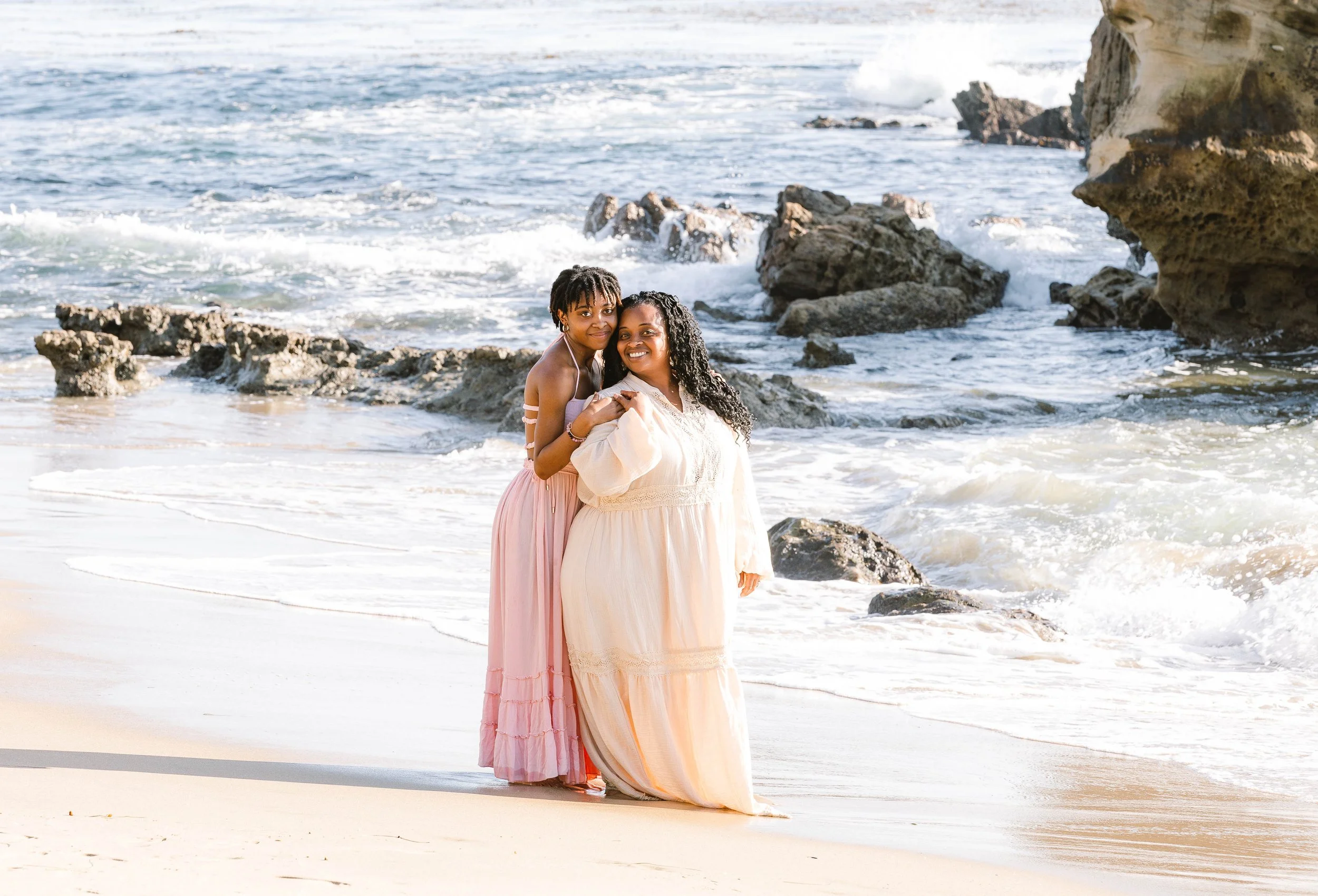 A mother and daughter standing on a sandy beach near the ocean with waves and rocks in the background, both smiling and wearing long flowing dresses.
