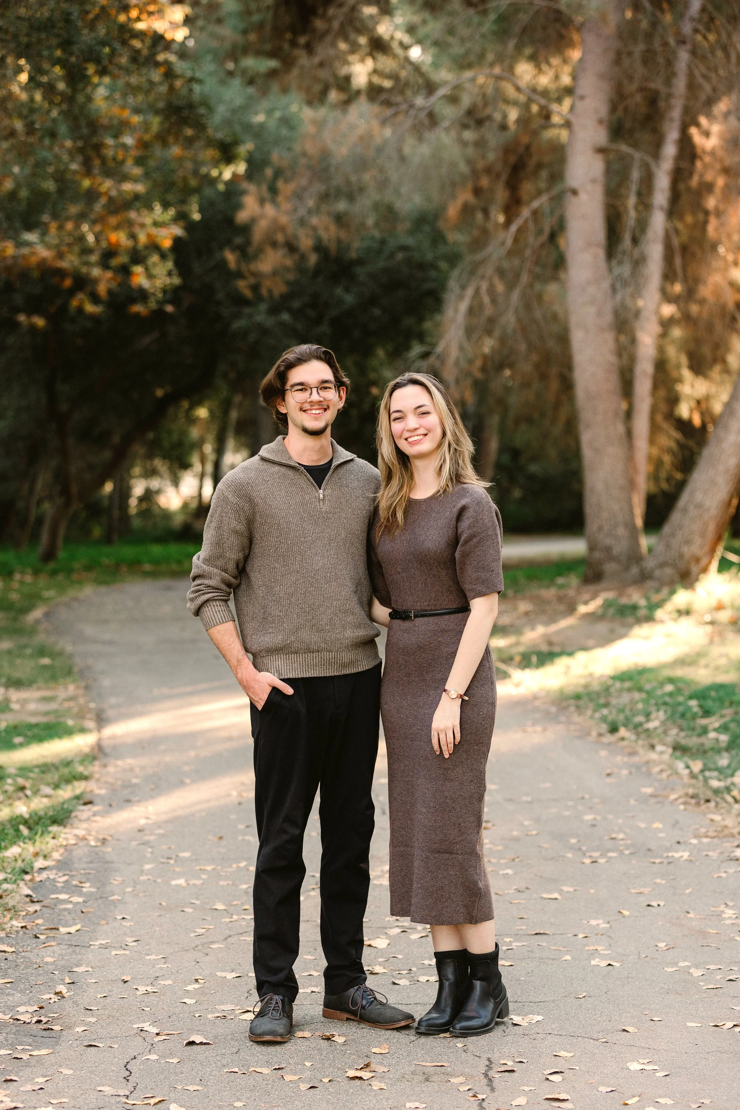 A smiling young man and woman standing close together outdoors on a path with fallen leaves, surrounded by trees with fall foliage.