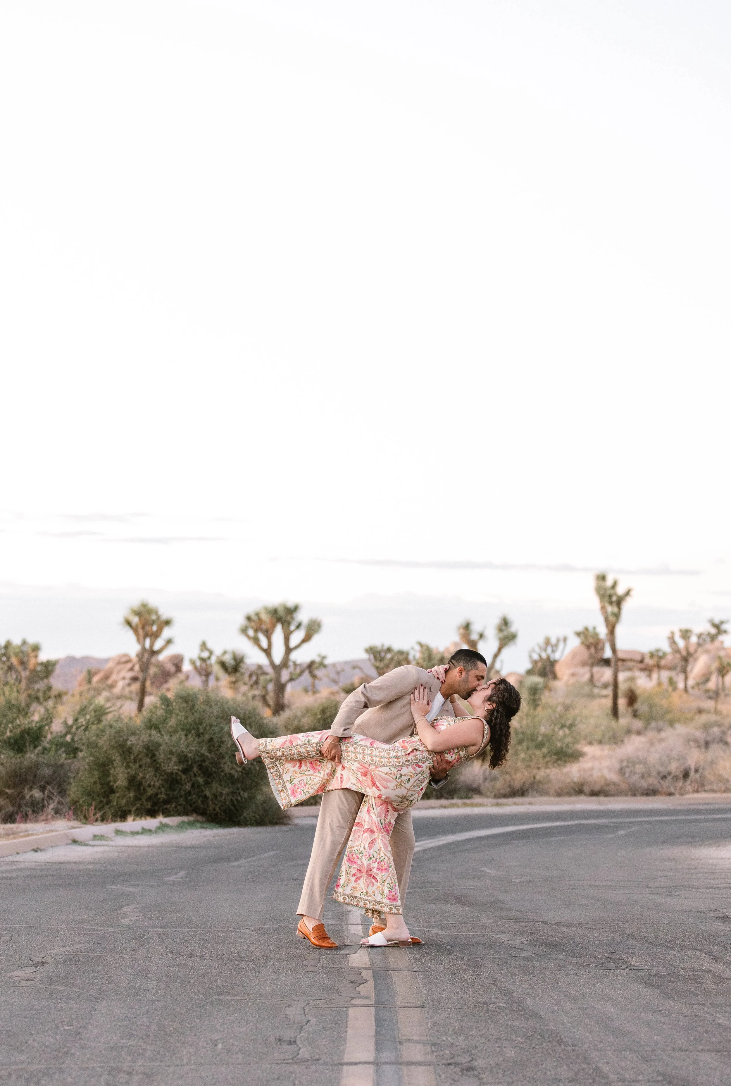 A man in a beige suit and a woman in a floral dress are standing on an empty road in a desert landscape, kissing, with the man holding the woman in his arms as she leans back.