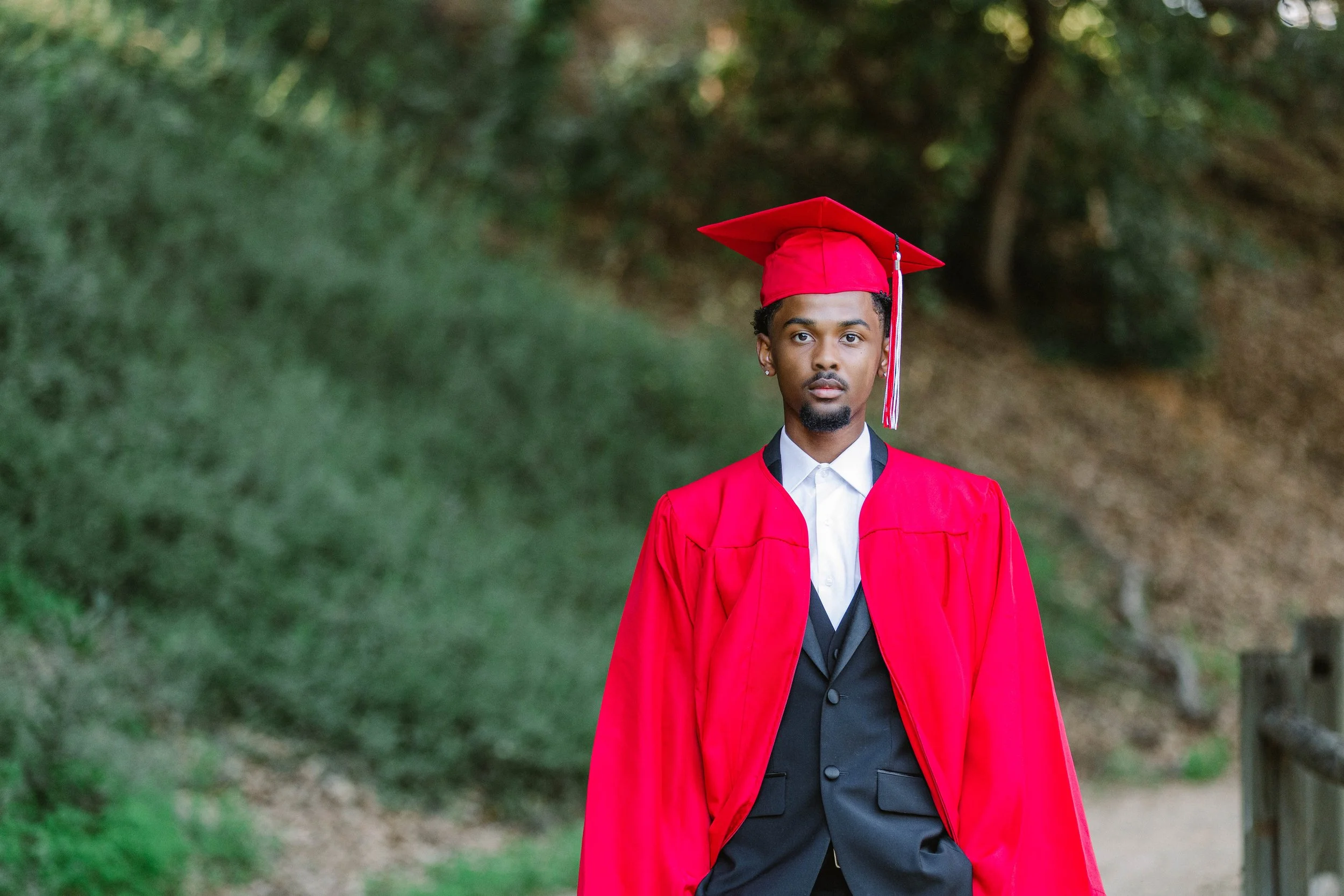A young man in a graduation cap and gown standing outdoors on a pathway with greenery in the background.